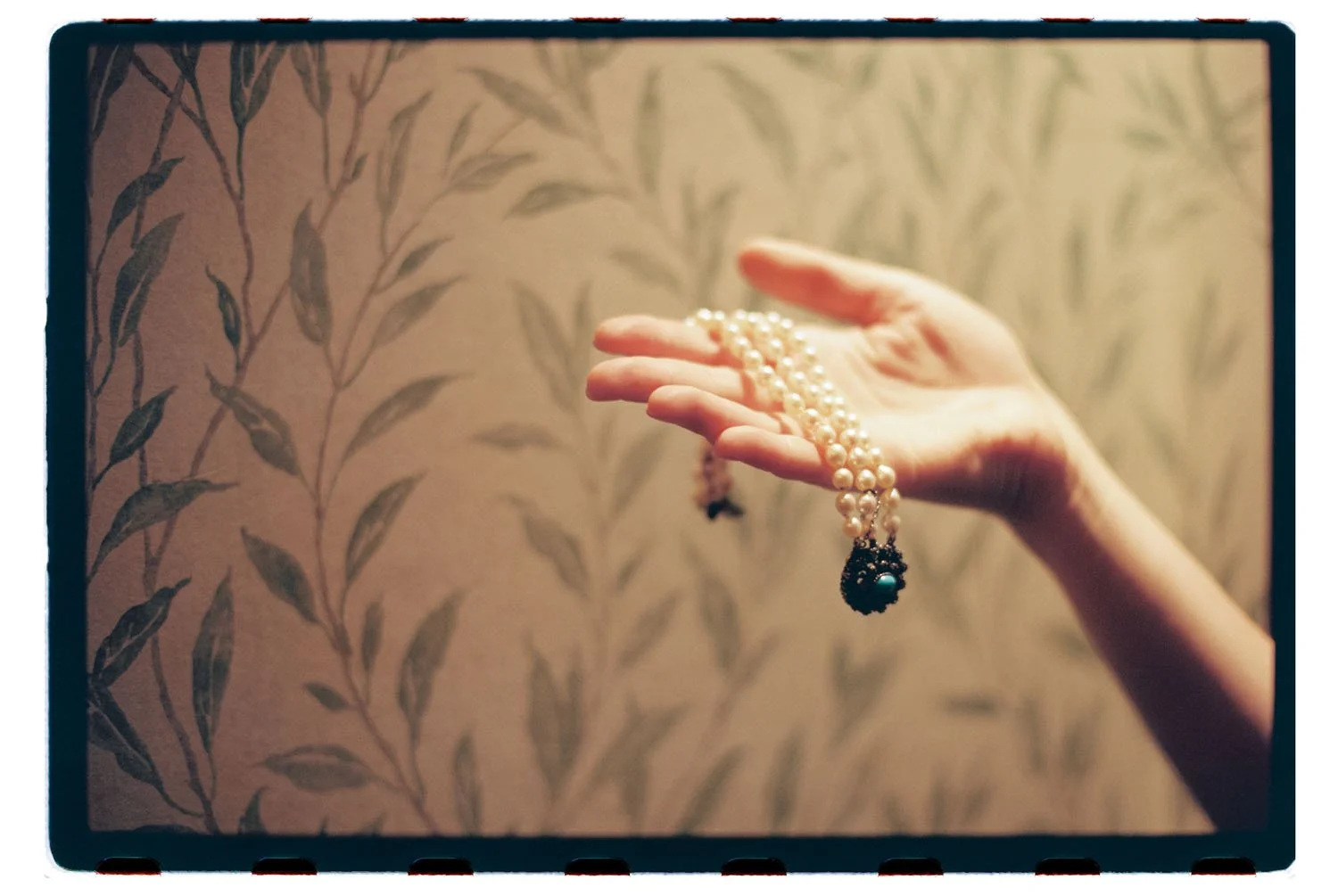 A hand holding a pearl necklace with a black and turquoise pendant against a background with a leafy pattern.