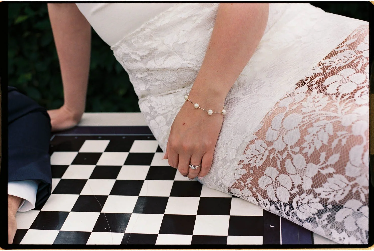 Close-up of a woman's hand with wedding ring and pearl bracelet resting on a black-and-white chessboard, wearing a white lace dress, with a man sitting nearby.