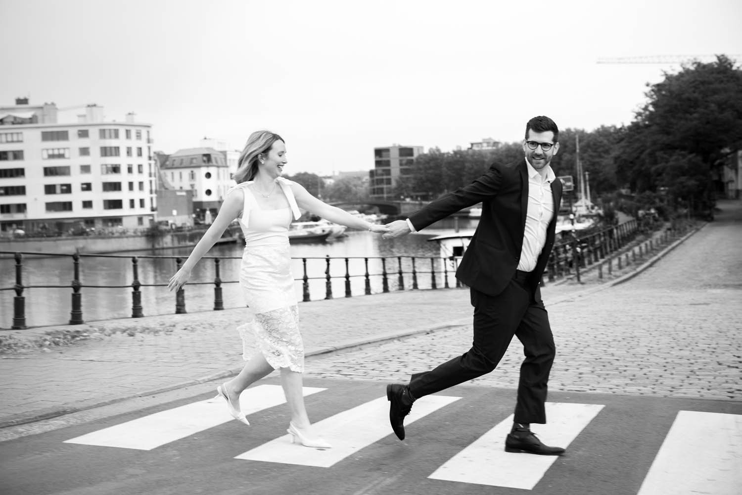 A black-and-white photo of a couple in formal attire holding hands and running across a pedestrian crosswalk near water and buildings, smiling and looking at each other.