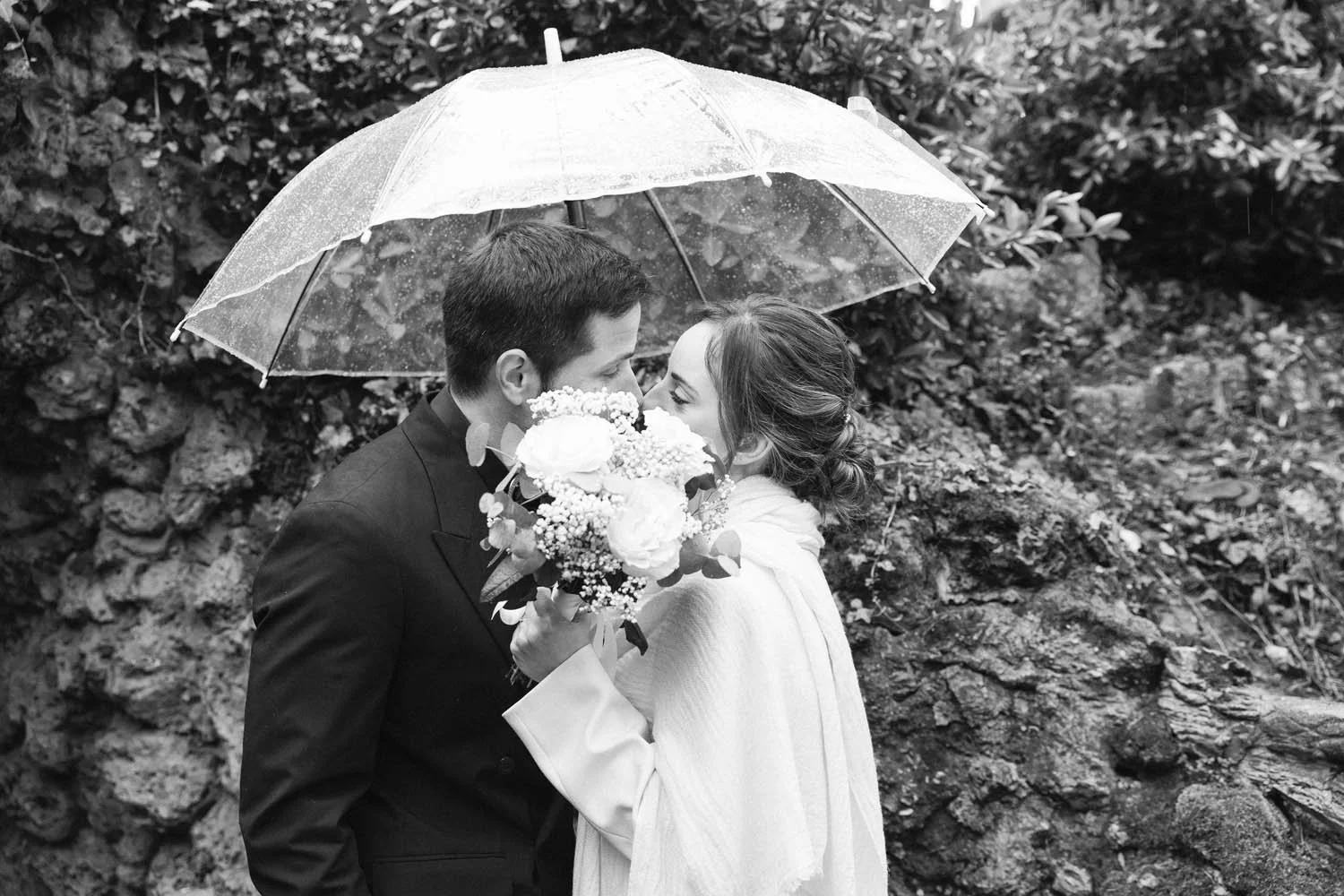 A black and white photo of a couple on their wedding day, standing close under a transparent umbrella in the rain, with the bride holding a bouquet of flowers.