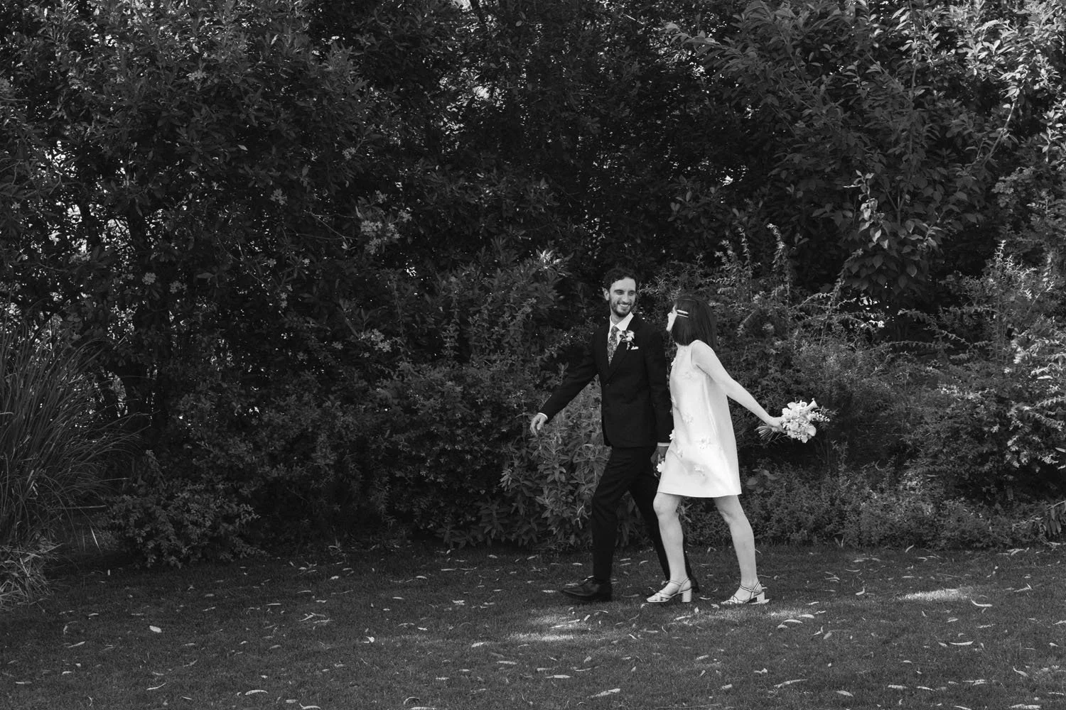 A black-and-white photo of a couple walking outdoors in front of dense bushes. The man, dressed in a tuxedo, is smiling and looking at the woman. The woman, wearing a light-colored dress and high heels, is holding a bouquet of flowers behind her back