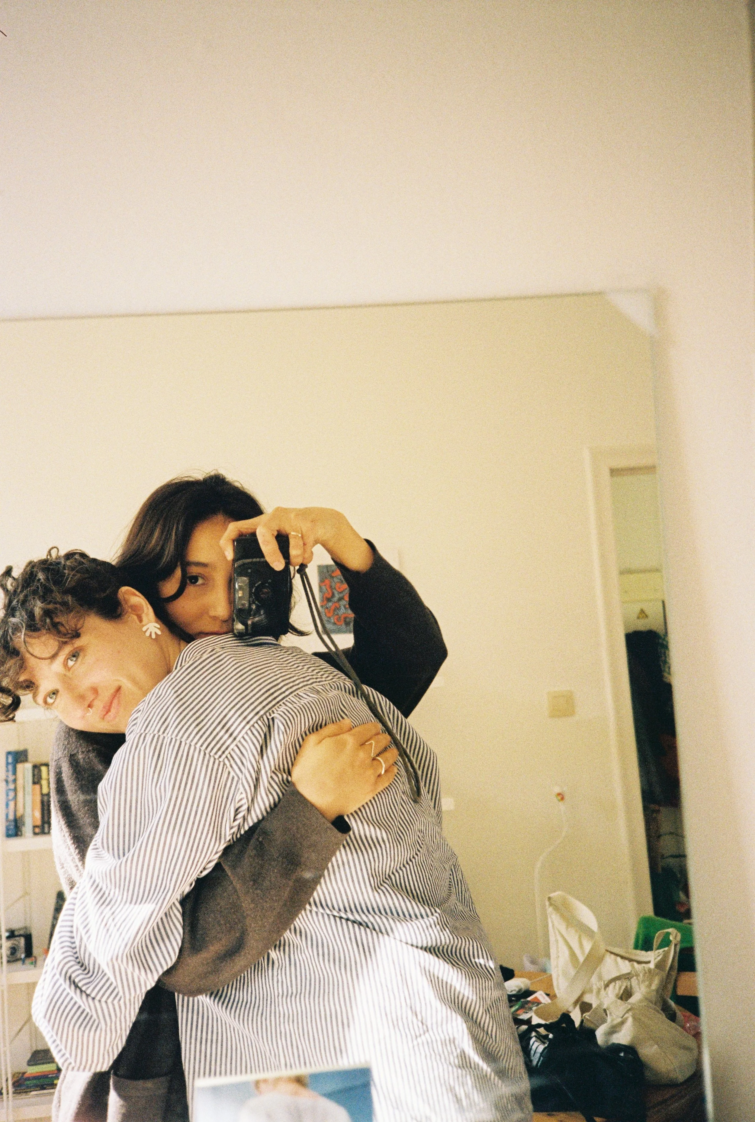 Two women taking a mirror selfie in a room. One woman with curly hair is smiling, and the other woman with dark straight hair is holding a camera, partially covering her face. There are bookshelves, a doorway, and various items on a table in the background.