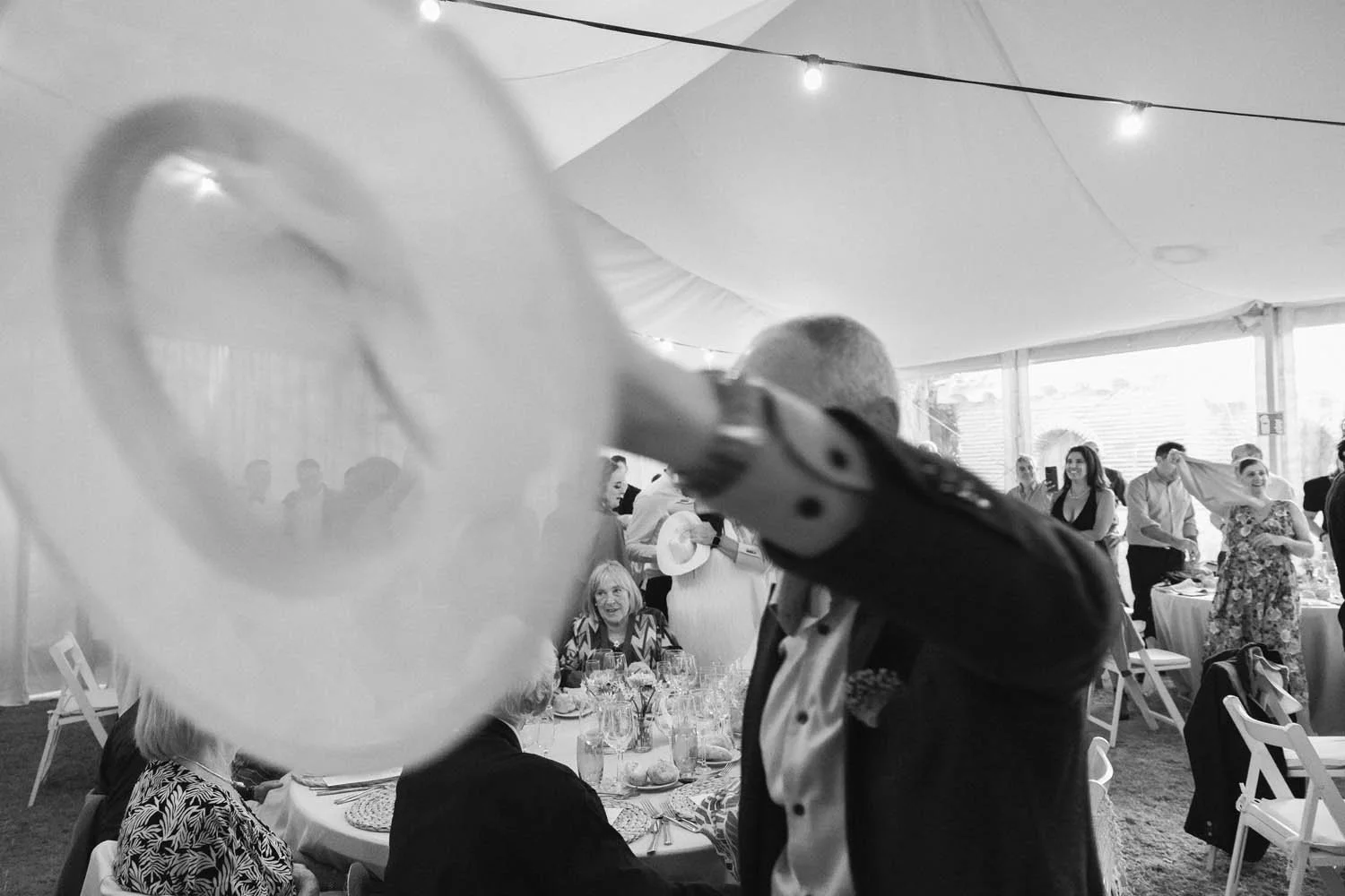 Documentary-style photography in black & white of a person holding a hat in a wedding celebration. They are dancing, and there are more people in the background.