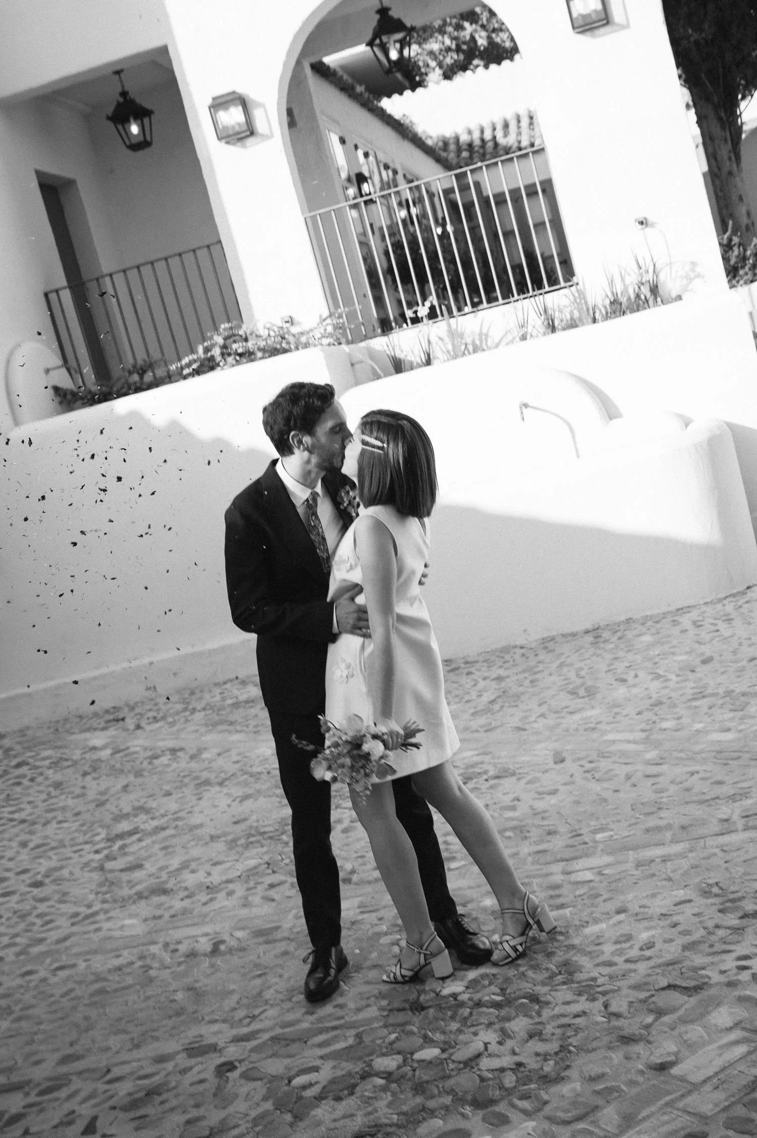 A black and white photo of a young couple kissing outdoors. The man is dressed in a suit and tie, and the woman is wearing a white dress and heels, holding a bouquet of flowers. They are standing on a cobblestone path with a white wall, plants, and l