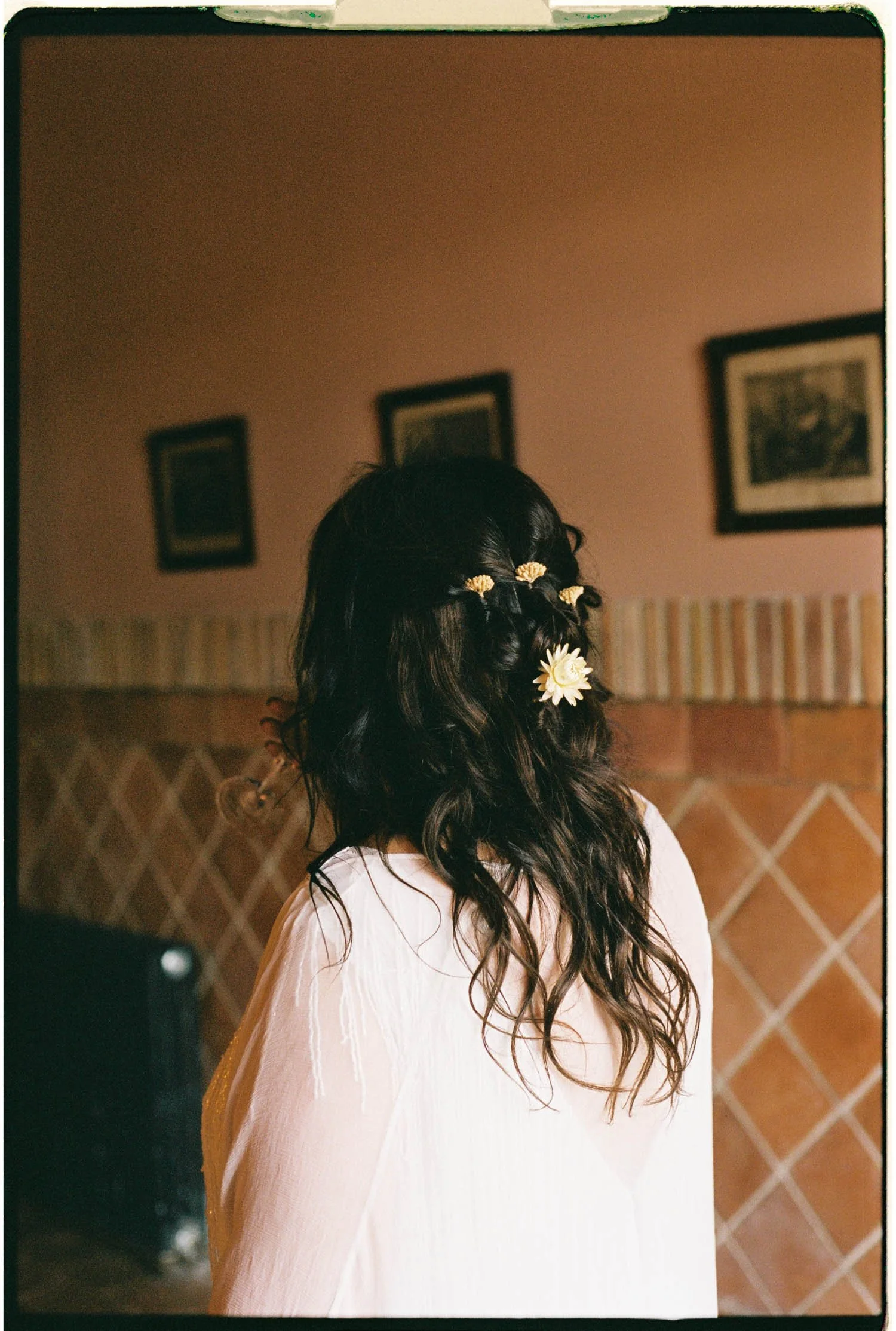 Back of a woman's head with dark, curly hair adorned with white and yellow flowers, wearing a light-colored, flowing dress.