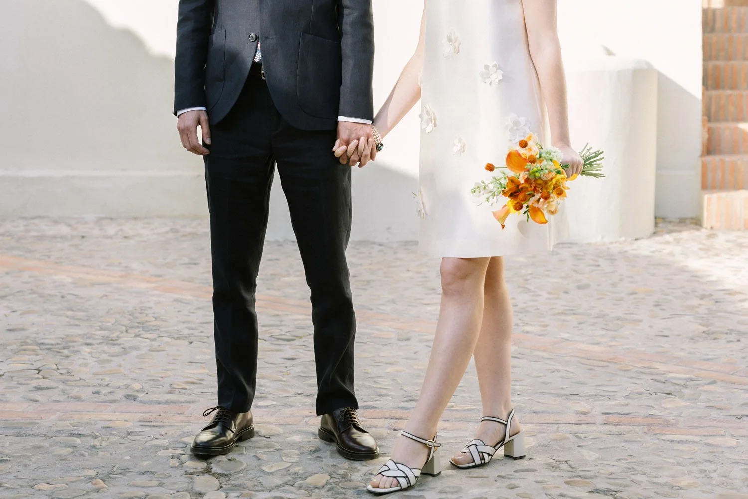 A couple holding hands, with the woman holding a bouquet of orange flowers, standing on a cobblestone street near white walls and brick steps.