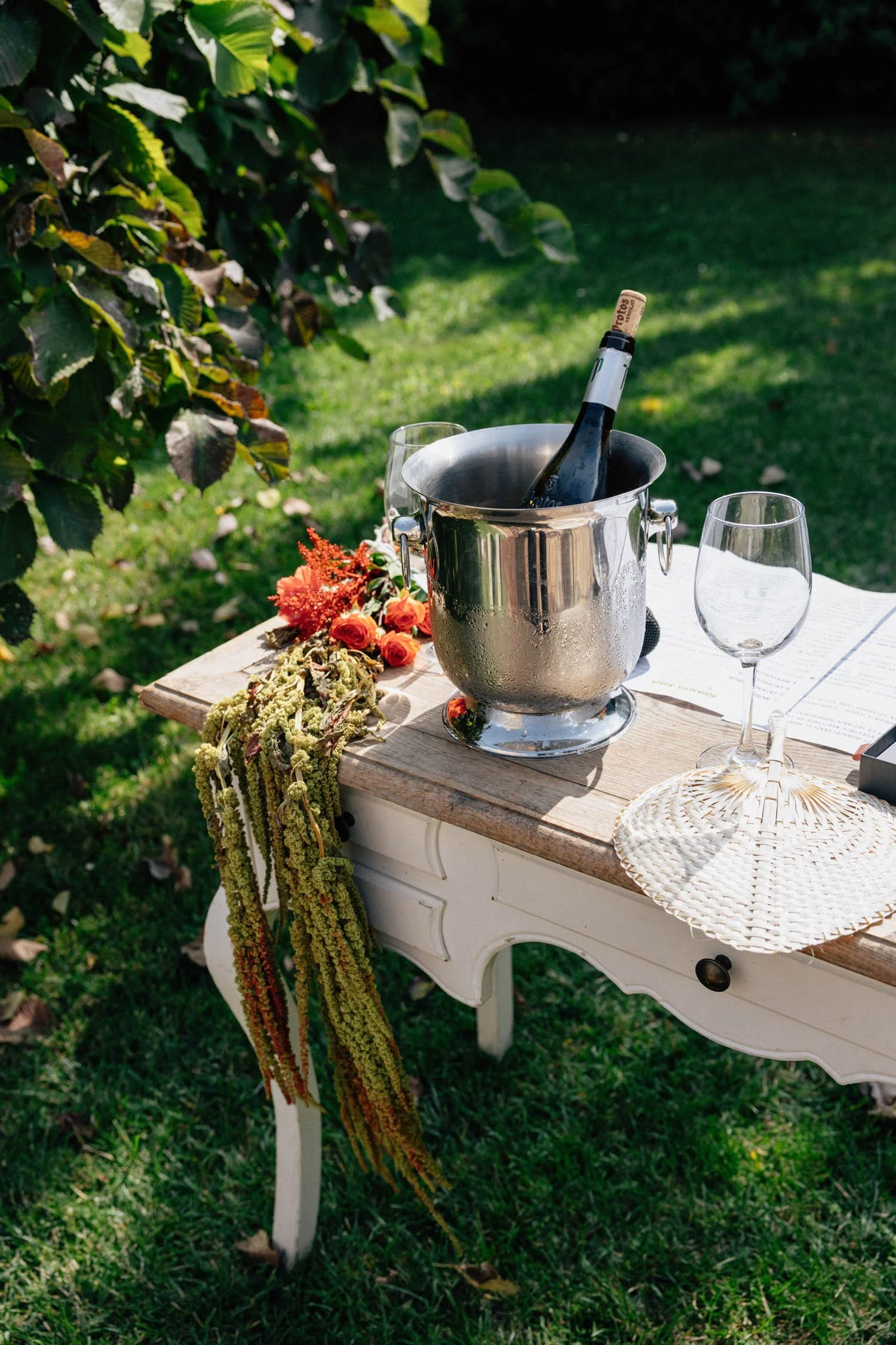 A white vintage side table outdoors on green grass, adorned with a bouquet of orange flowers and greens, an open book, a wicker coaster, an empty wine glass, and a silver ice bucket with a bottle of wine. There are trees and shady areas in the background.