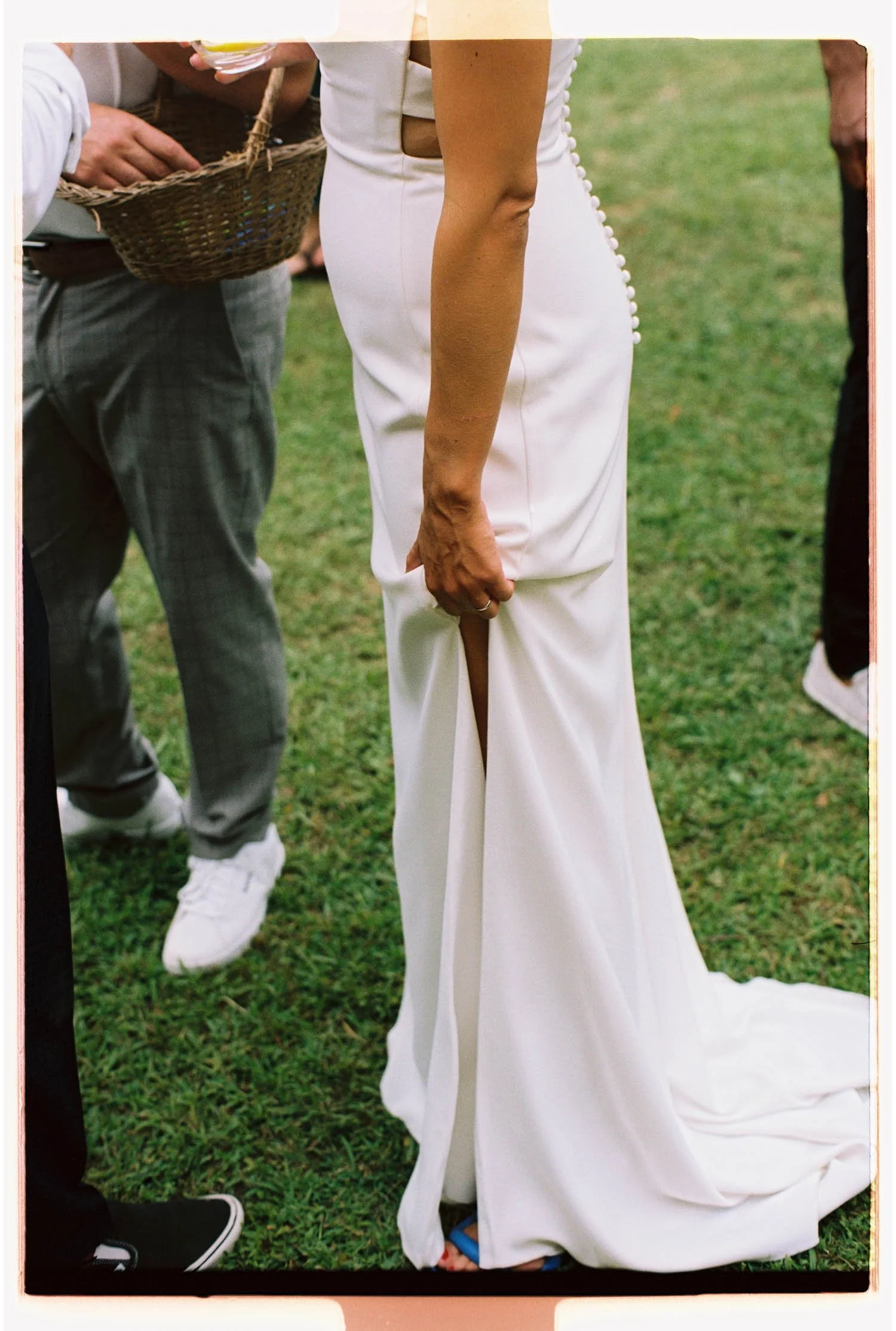 A woman in a white dress with pearl buttons standing on grass, with a person wearing gray pants and black sneakers nearby. Part of the woman’s dress is lifted slightly revealing her foot in a blue sandal.