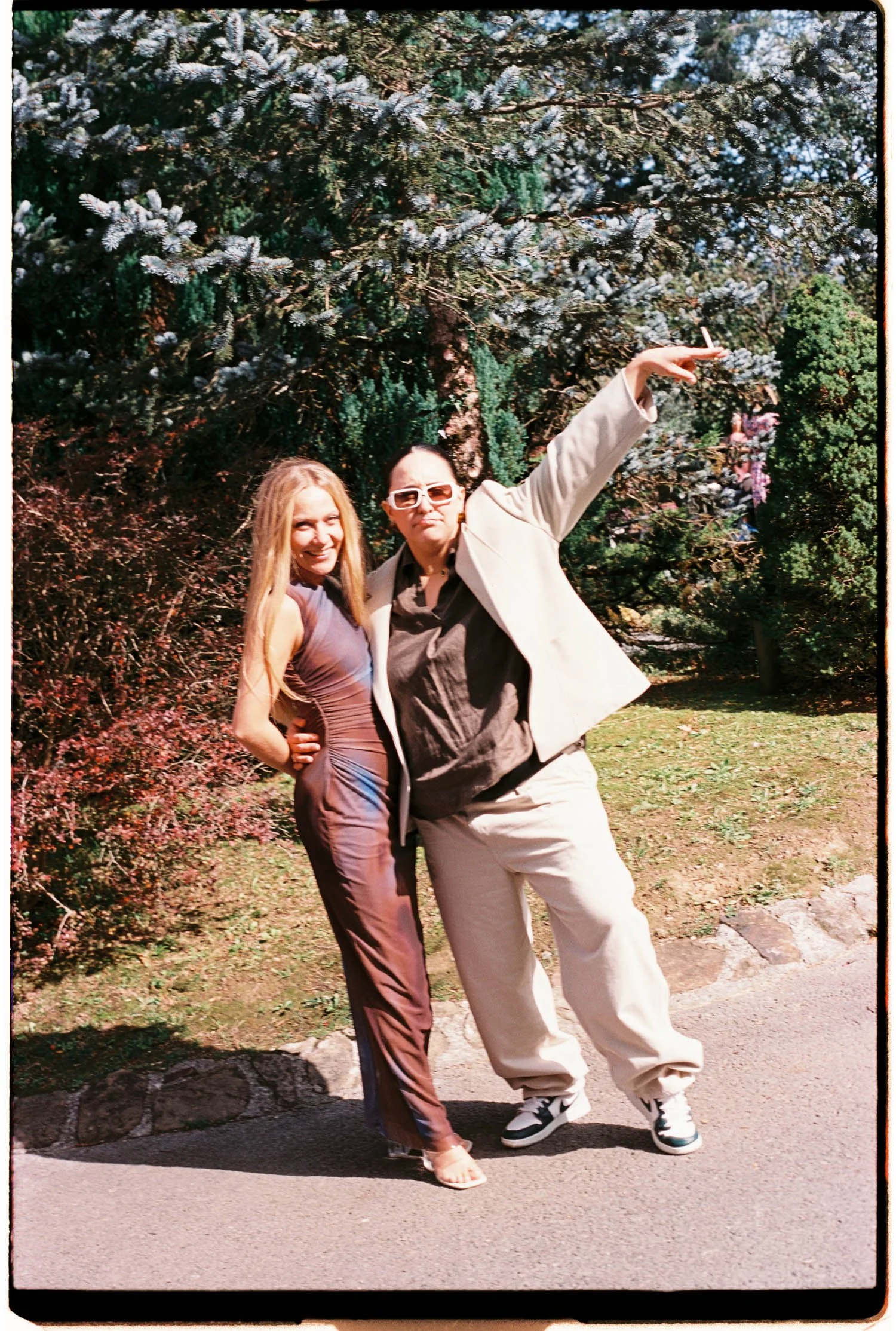 Two women posing outdoors in front of a large evergreen tree, one wearing a shiny dress and the other in a beige suit, with one woman holding a cigarette.