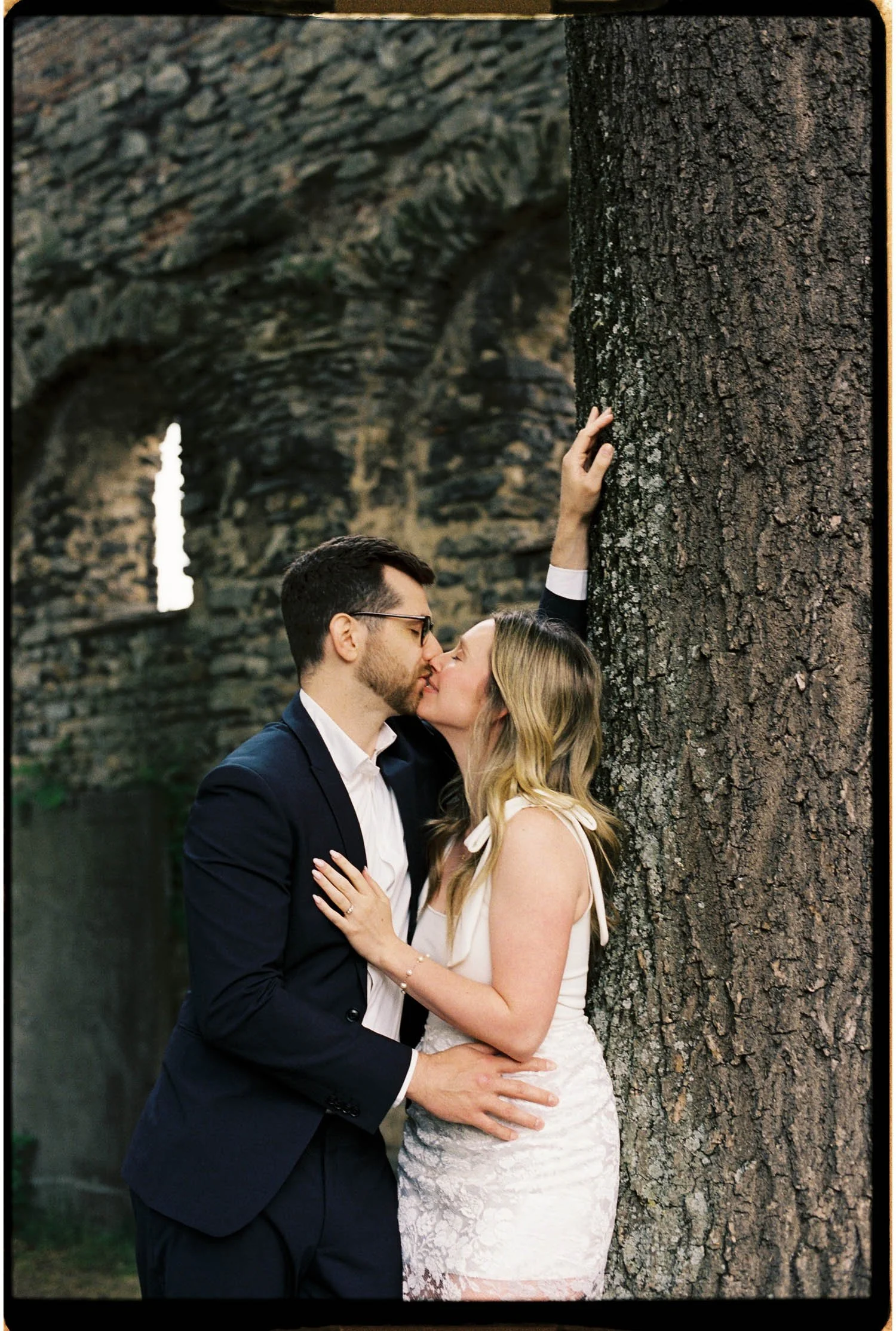 A couple before the wedding ceremony kissing outdoors by a large tree, with the woman’s hand on the tree and her other hand on the man’s neck, in front of a stone archway.
