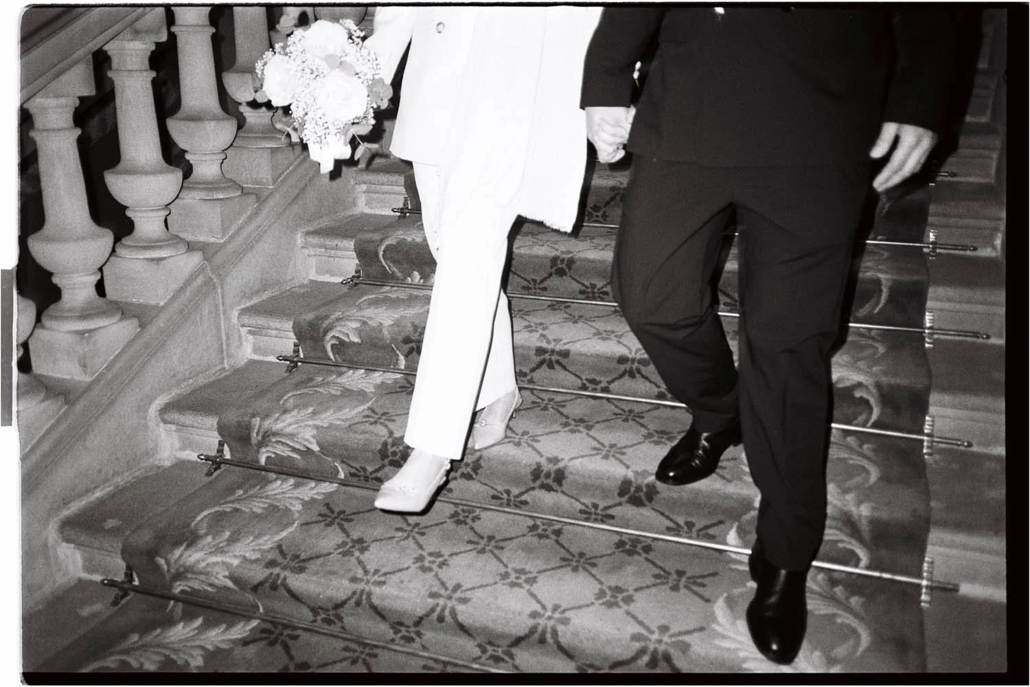 Analogue black & white photo of a just-married couple holding hands, walking up a carpeted staircase with floral patterns, with a bride holding a bouquet visible at the top of the stairs.
