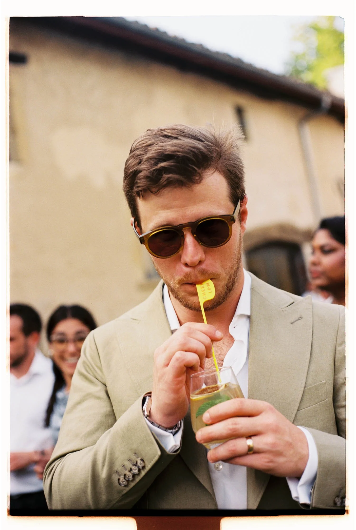 Man in beige suit and sunglasses holding a drink with a yellow straw at a social gathering, with a woman and other people in the background.