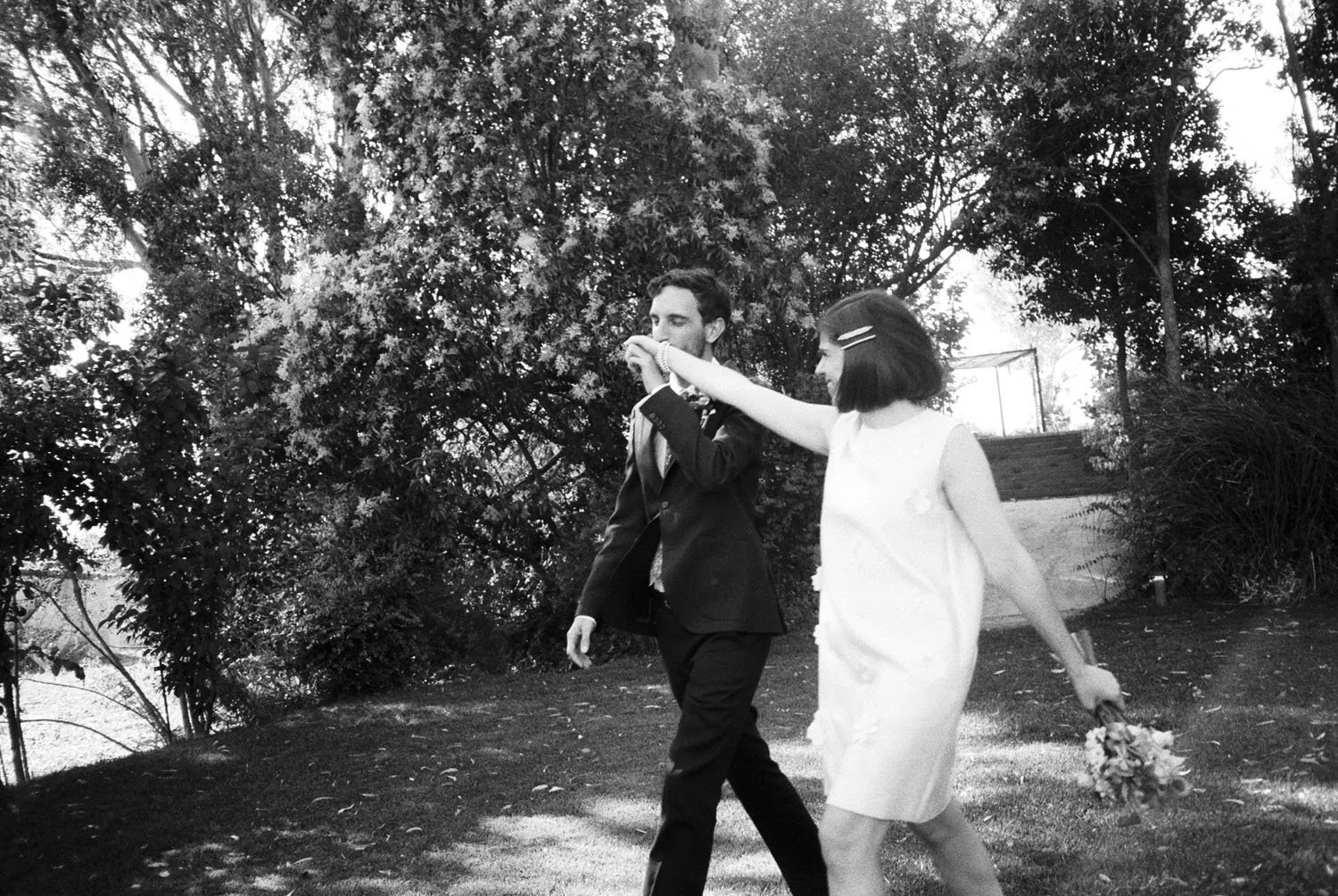 A black-and-white photo of a couple walking outdoors, with trees in the background. The woman is holding a bouquet in one hand and is being kissed by the groom in the other hand.