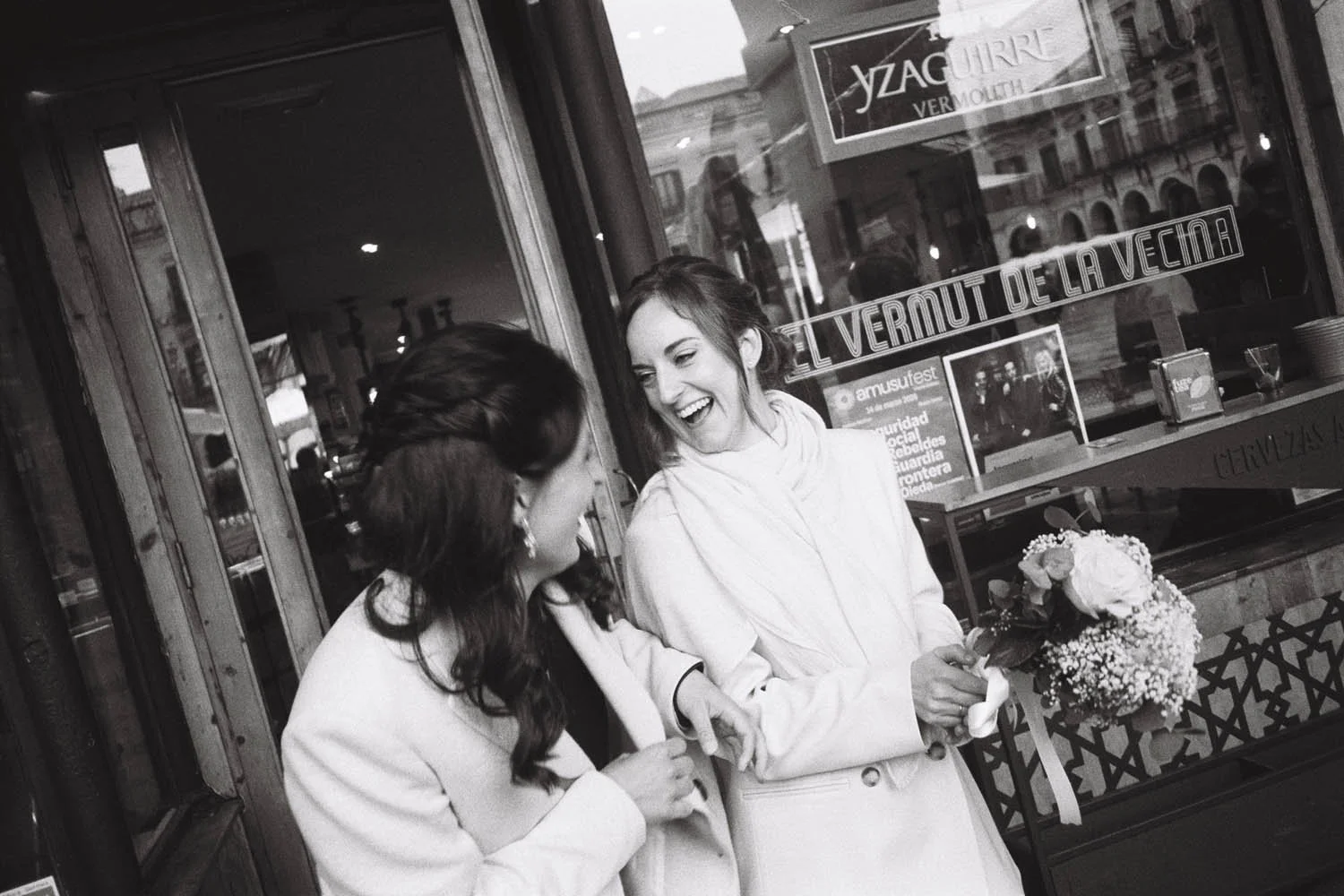 Two women smiling and laughing outside a restaurant, one holding a bouquet of flowers, both dressed in white, engaging in conversation.