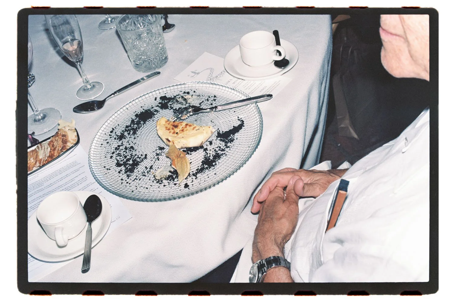 Analogue photo with flash of a wedding dinner table with a partially eaten slice of pie on a glass plate, surrounded by used utensils, drinks, and menus, with a person in a white chef's coat sitting nearby.