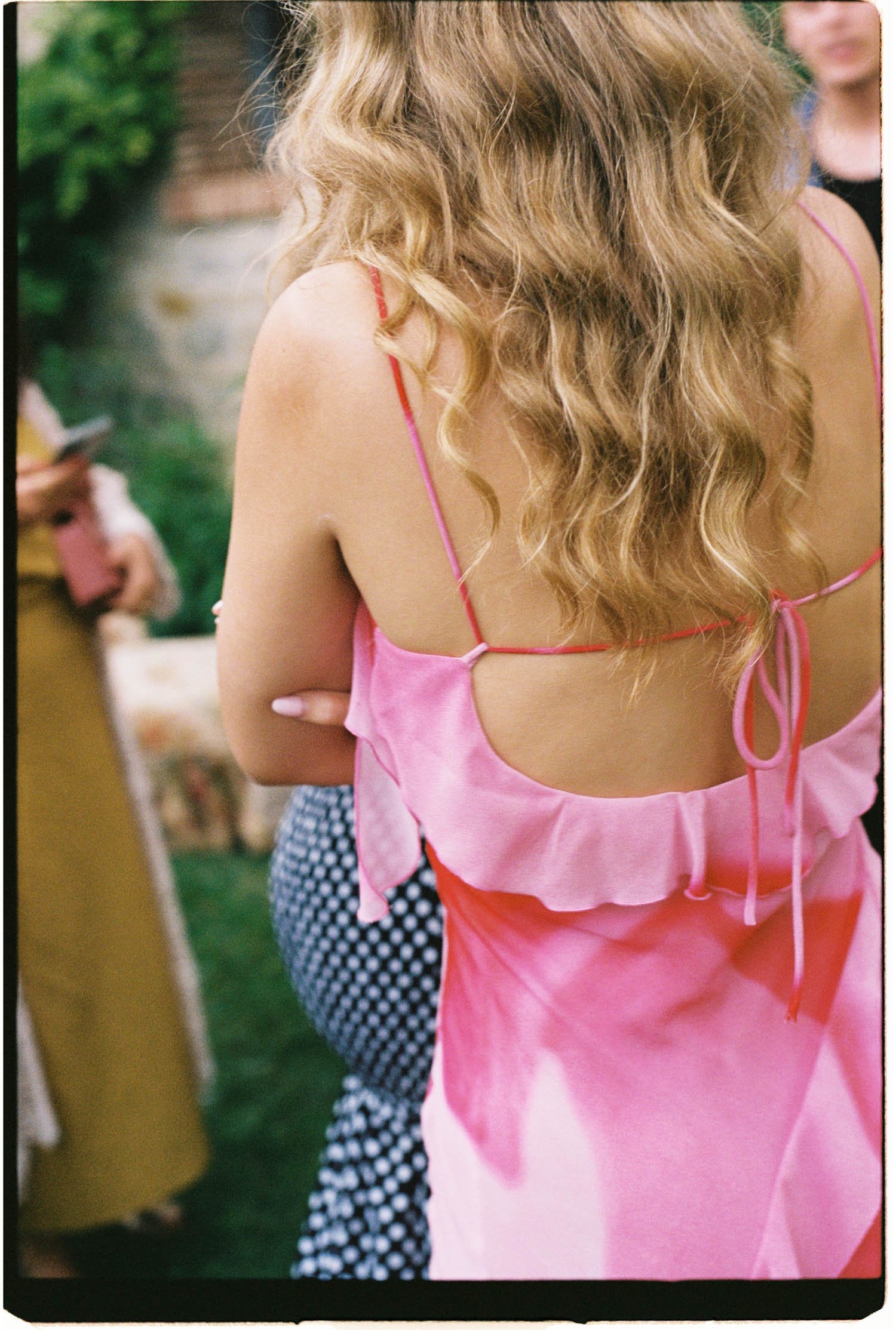 Close-up of the back of a wedding guest with wavy blonde hair wearing a pink sleeveless dress, standing outdoors.