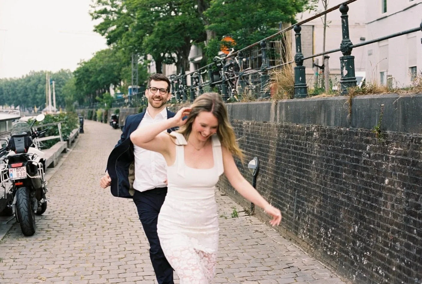 A couple in wedding clothes walking along a cobblestone path by a canal, smiling and laughing, with parked bicycles and trees in the background.