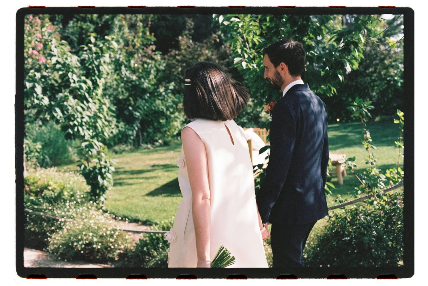 A couple holds hands outdoors in a garden, dressed in wedding attire, with the woman in a white dress holding a bouquet and the man in a dark suit.