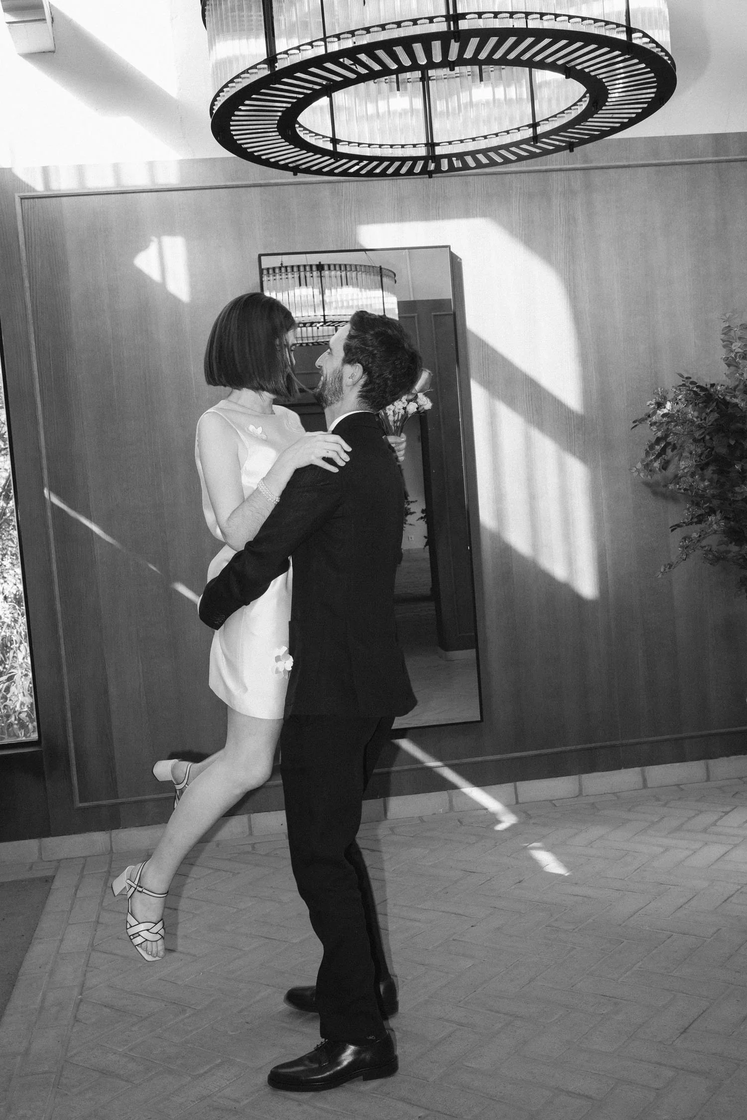 A couple dancing in front of a mirror in a luxury room. The woman is wearing a white dress and high-heeled shoes, while the man is dressed in a dark suit. The sunlight creating shadows on the wall.