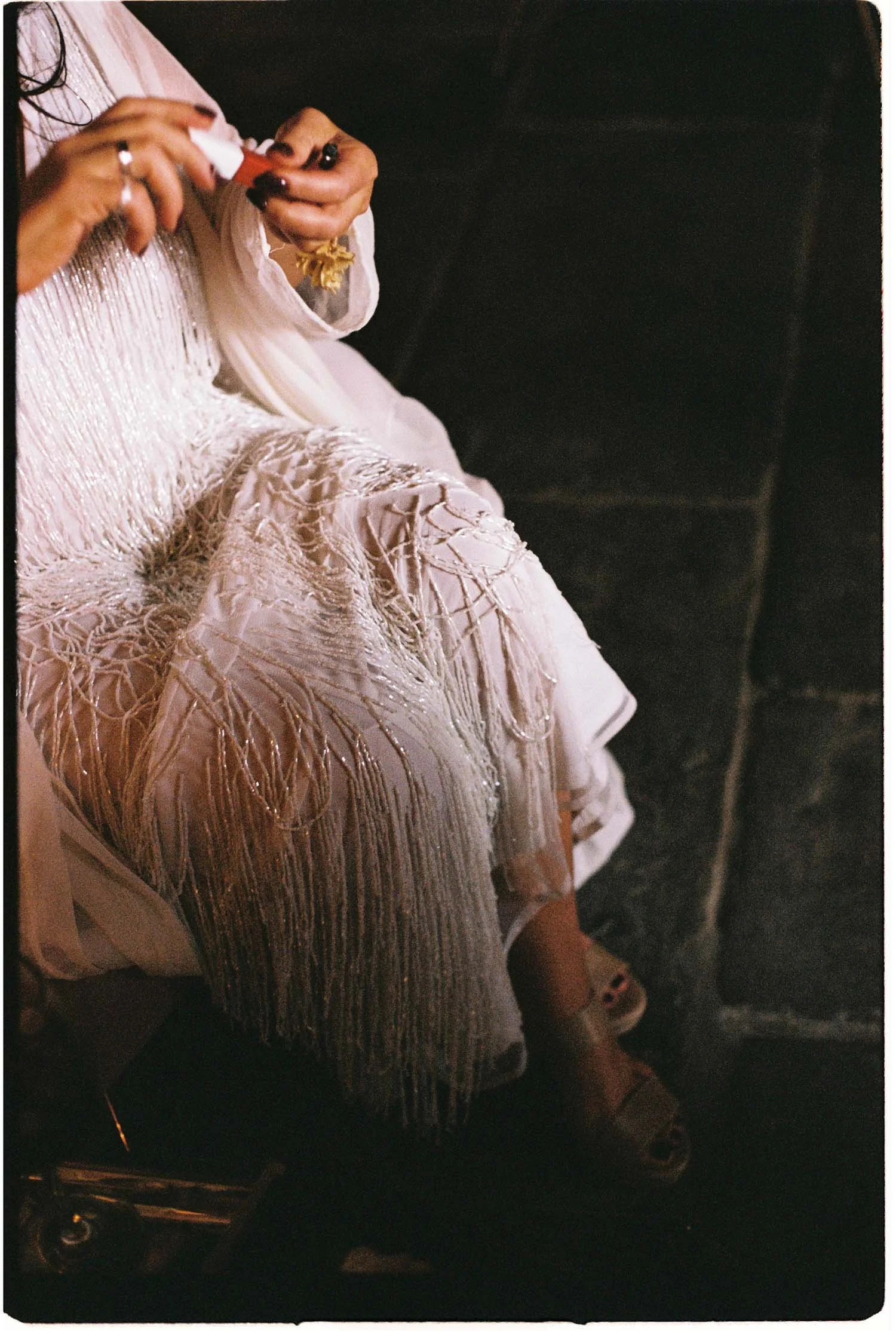 The bride is wearing an elegant, white, embroidered dress with intricate gold threadwork and bead detailing, sitting on a chair in a dimly lit setting.