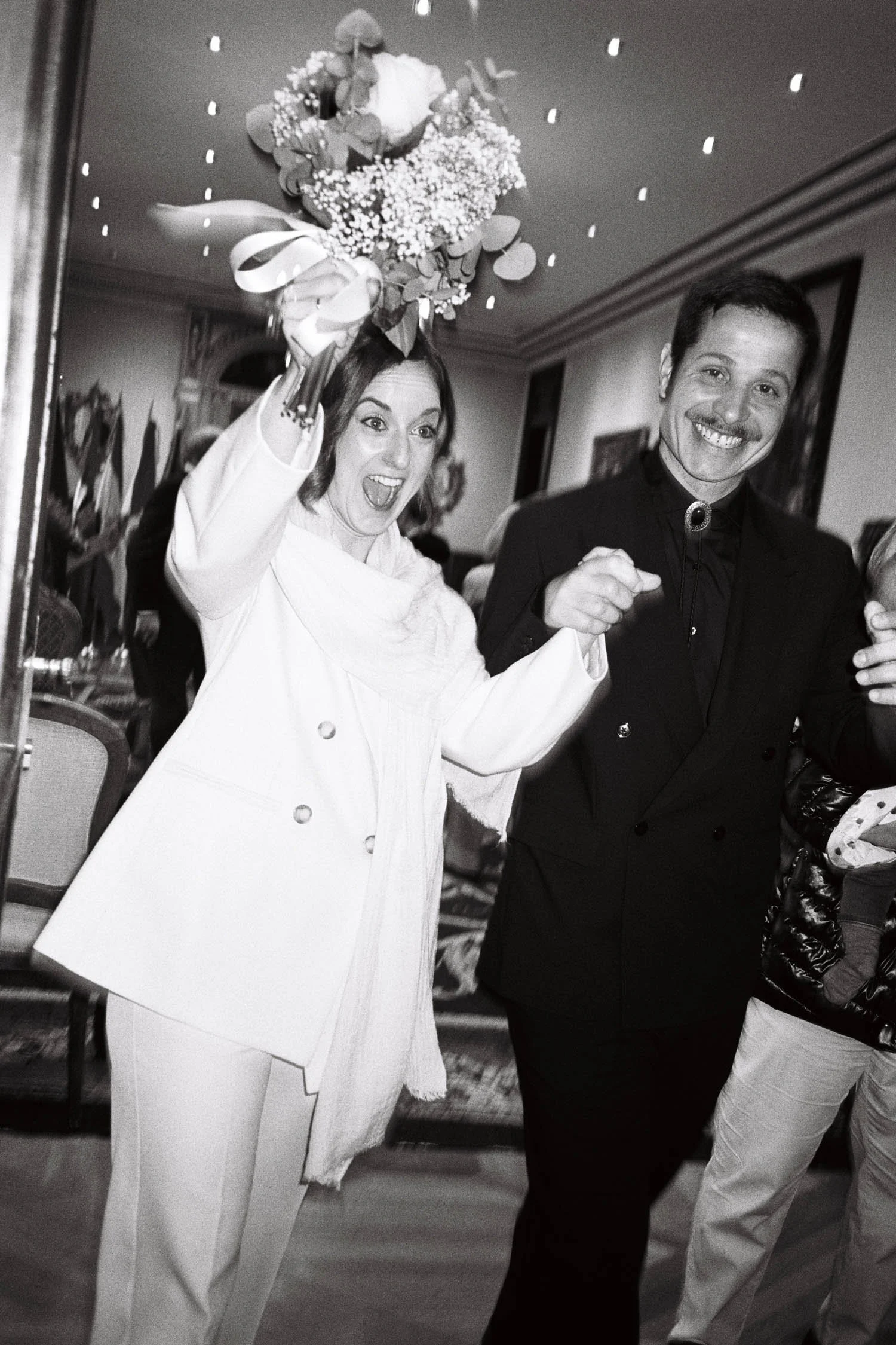 A couple smiling, celebrating their wedding. Holding a bouquet above her head and wearing a white suit. The man is wearing a dark modern suit and is making a fist gesture. The background shows a civil wedding room.