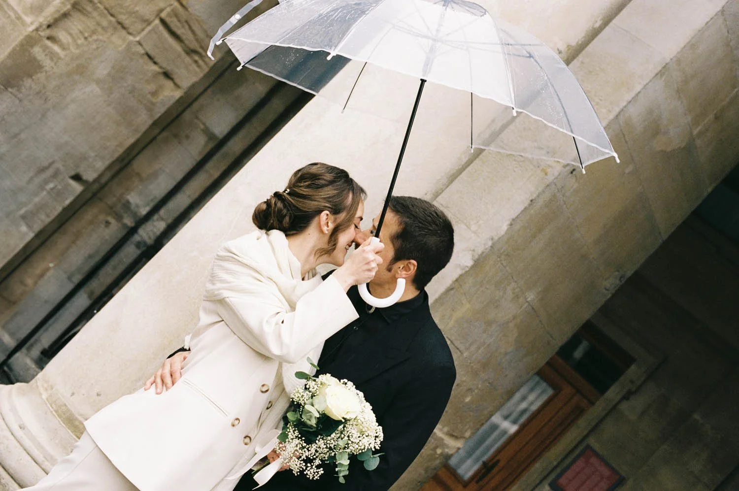 A couple sharing a romantic moment under an umbrella outside after the wedding ceremony, with the woman holding a bouquet of white flowers.
