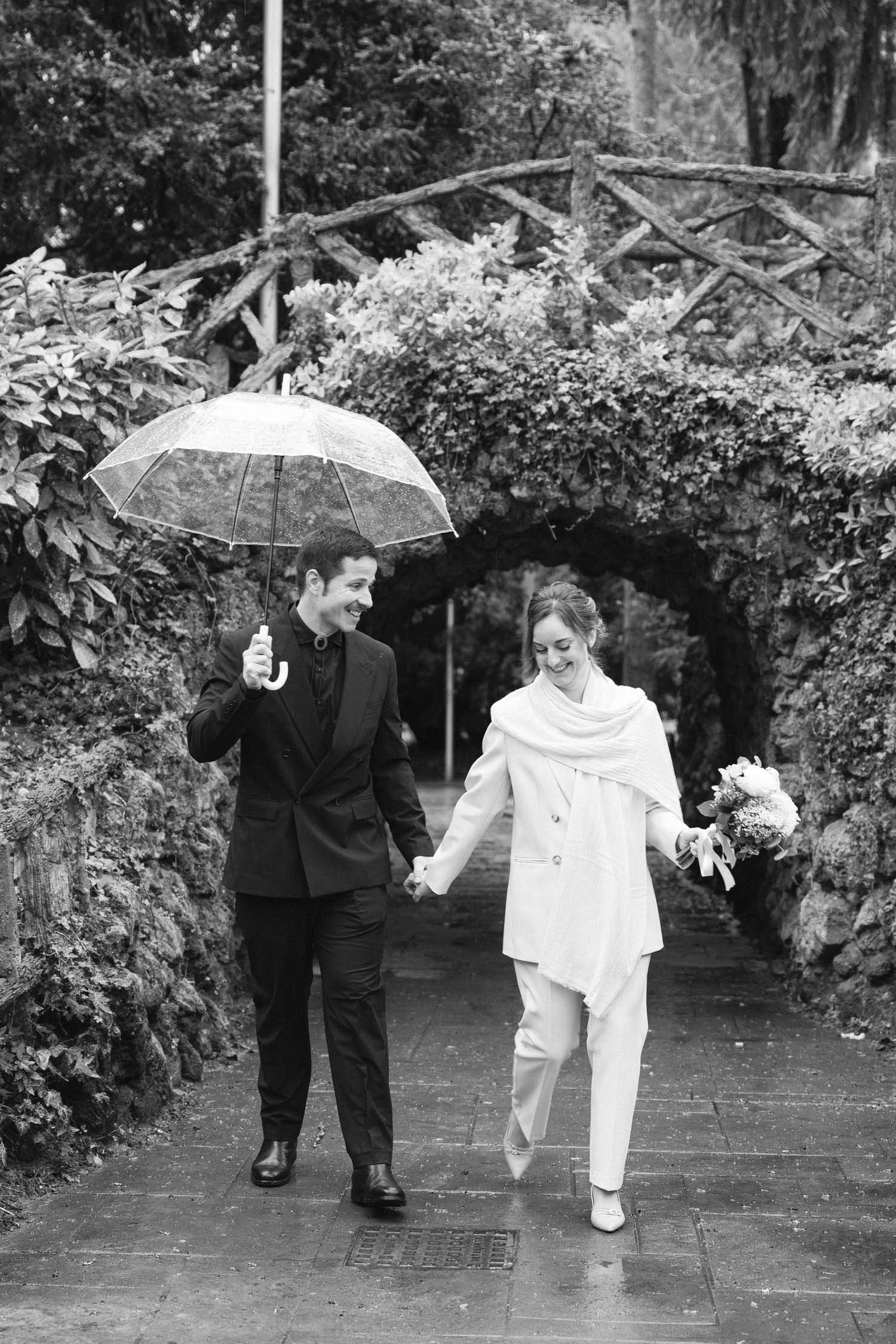 A black and white photo of a smiling couple walking hand in hand outdoors on a rainy day. The man is holding an umbrella and wearing a suit, while the woman is dressed in a coat and pants, holding a bouquet of flowers.