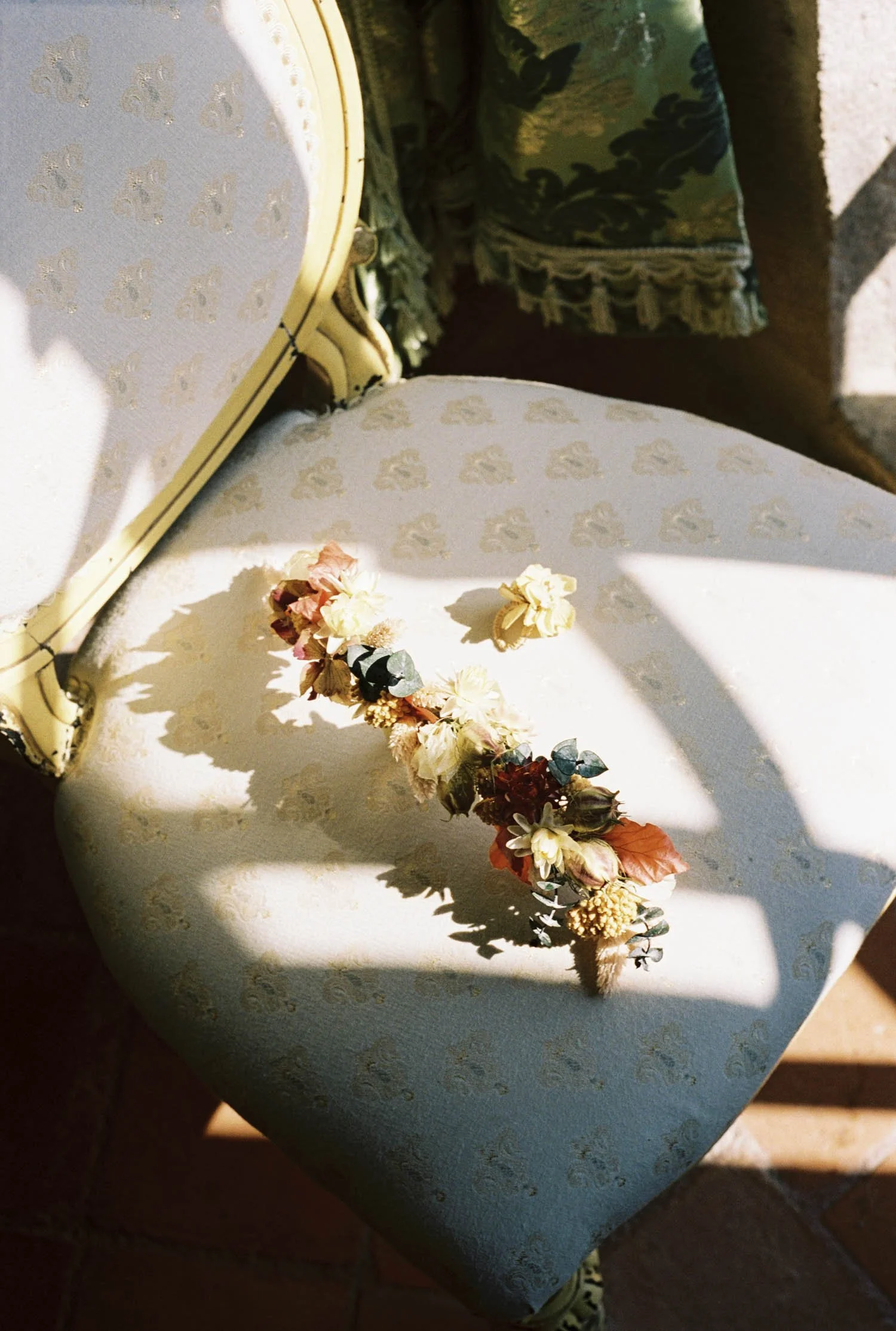 A floral crown with pink, yellow, white, and orange flowers resting on a white upholstered chair with gold trim, beside another similar chair, on a patterned fabric with tassel fringe in the background.