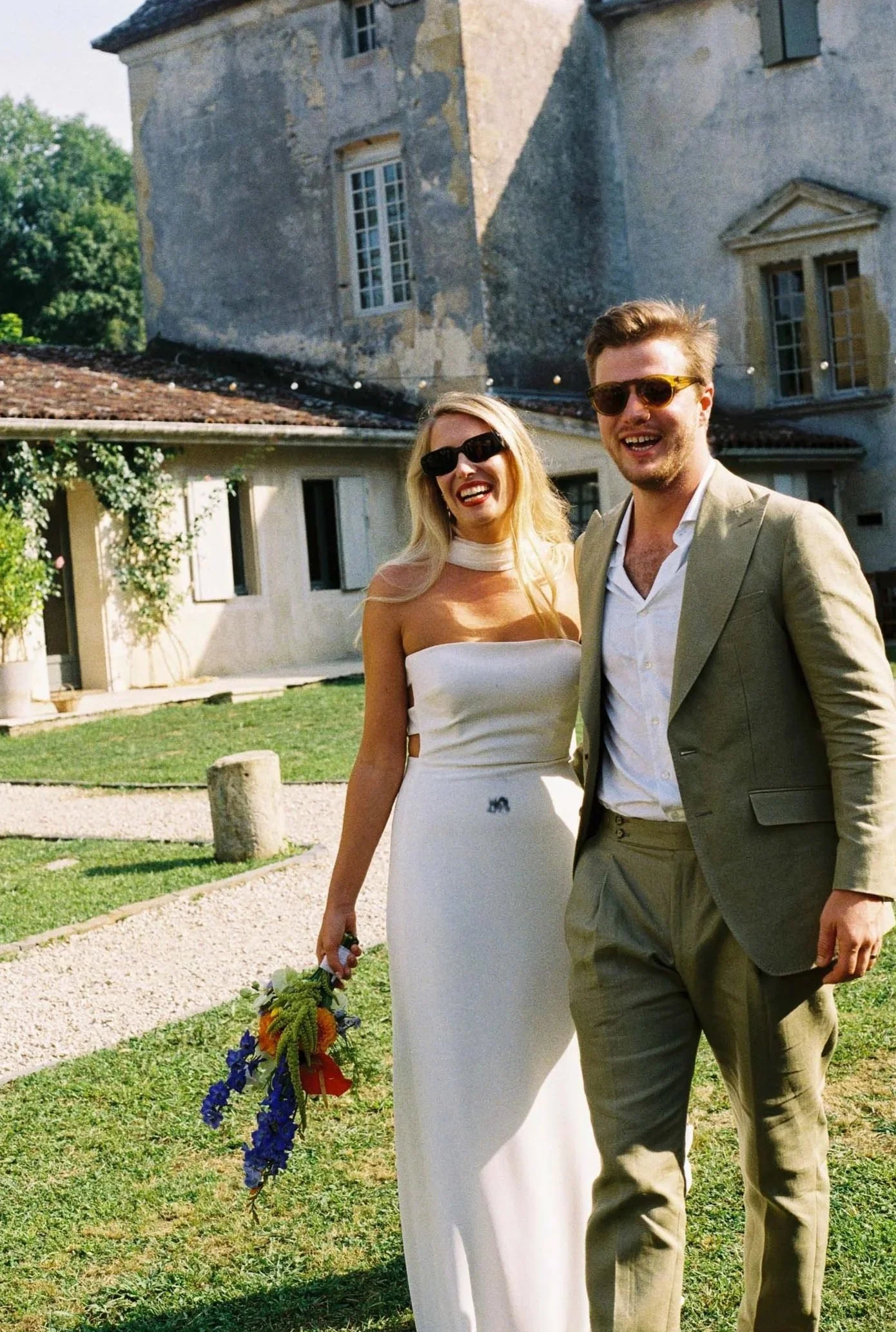 A smiling couple dressed in wedding attire walking outdoors in front of an old stone building with large windows and a tiled roof. The woman is holding a colorful bouquet of flowers, and both are wearing sunglasses.