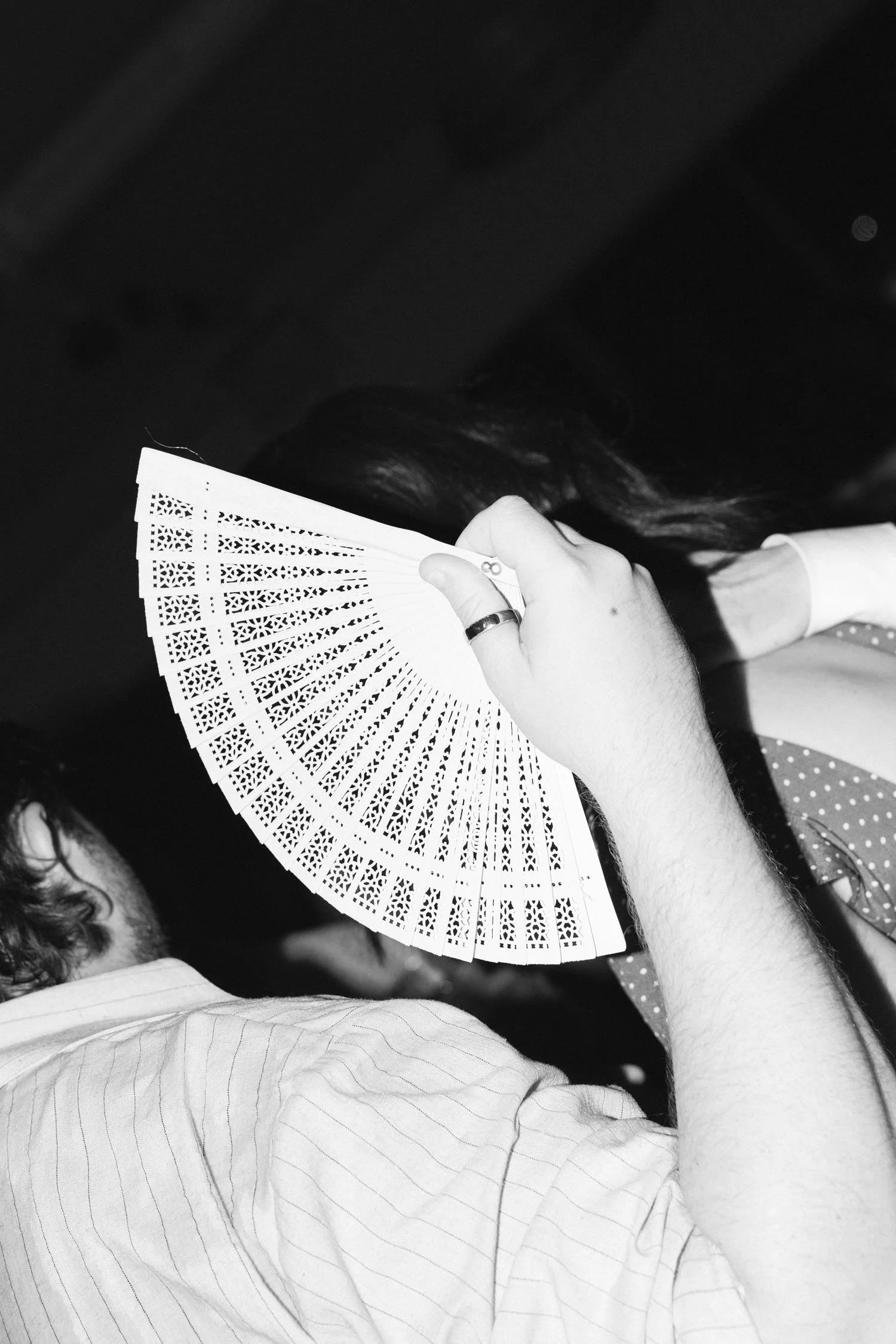 Person in a wedding party holding a decorative fan over their face, wearing a striped shirt, in a black and white photograph.