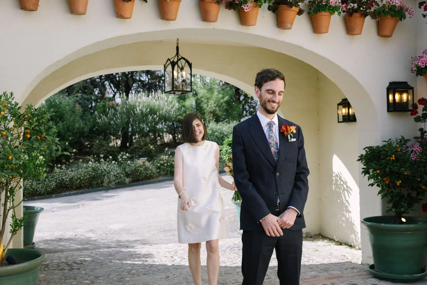 A smiling man in a dark suit with a colorful tie and flower boutonniere stands outside under an archway with potted plants and lanterns. A woman in a white dress with floral embroidery approaches him, smiling and holding a bouquet of flowers.