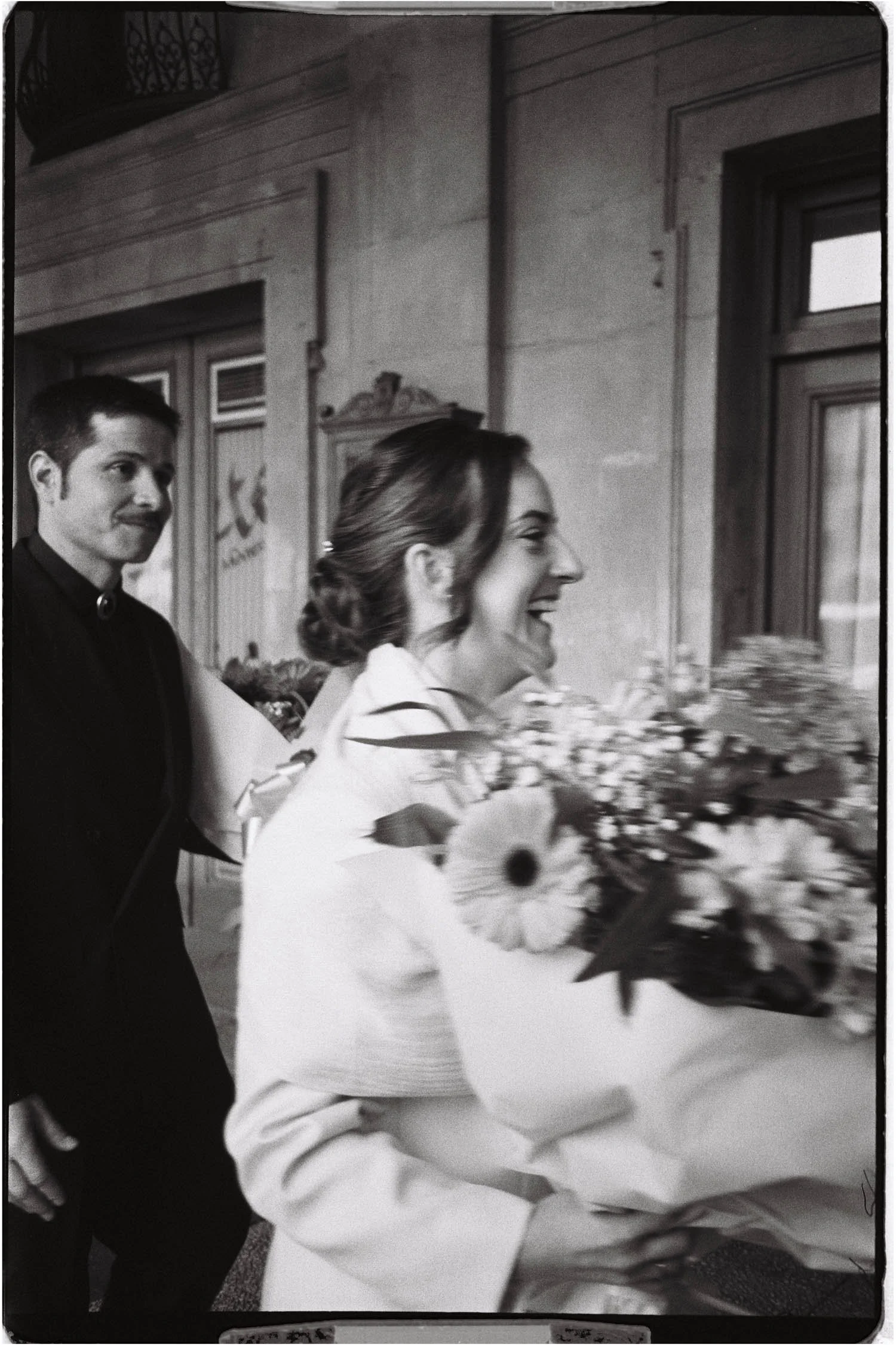 A woman in a white dress receiving a bouquet of flowers from a woman in a white dress. A man in a black suit stands nearby. The scene appears to take place indoors with wooden walls and windows.