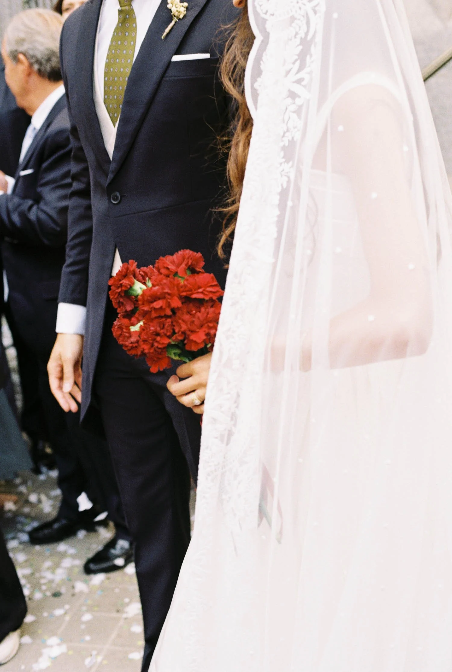 A bride and groom stand side by side at a wedding ceremony, holding hands. The bride wears a white dress and lace veil, while the groom wears a dark suit with a white shirt, green polka-dot tie, and a boutonniere. The bride holds a bouquet of red flo
