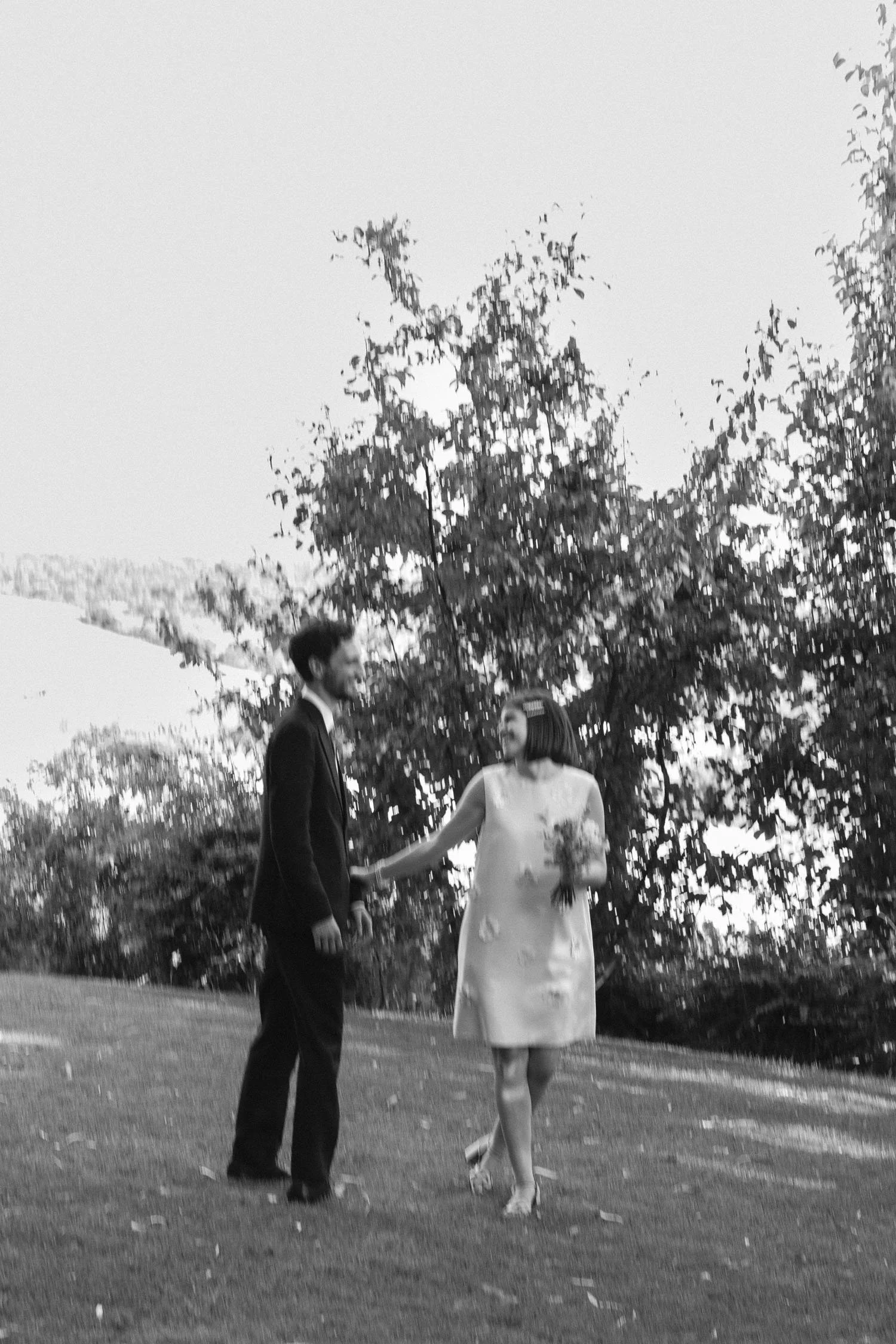 A couple walking outdoors holding hands, with trees and a grassy area in the background.