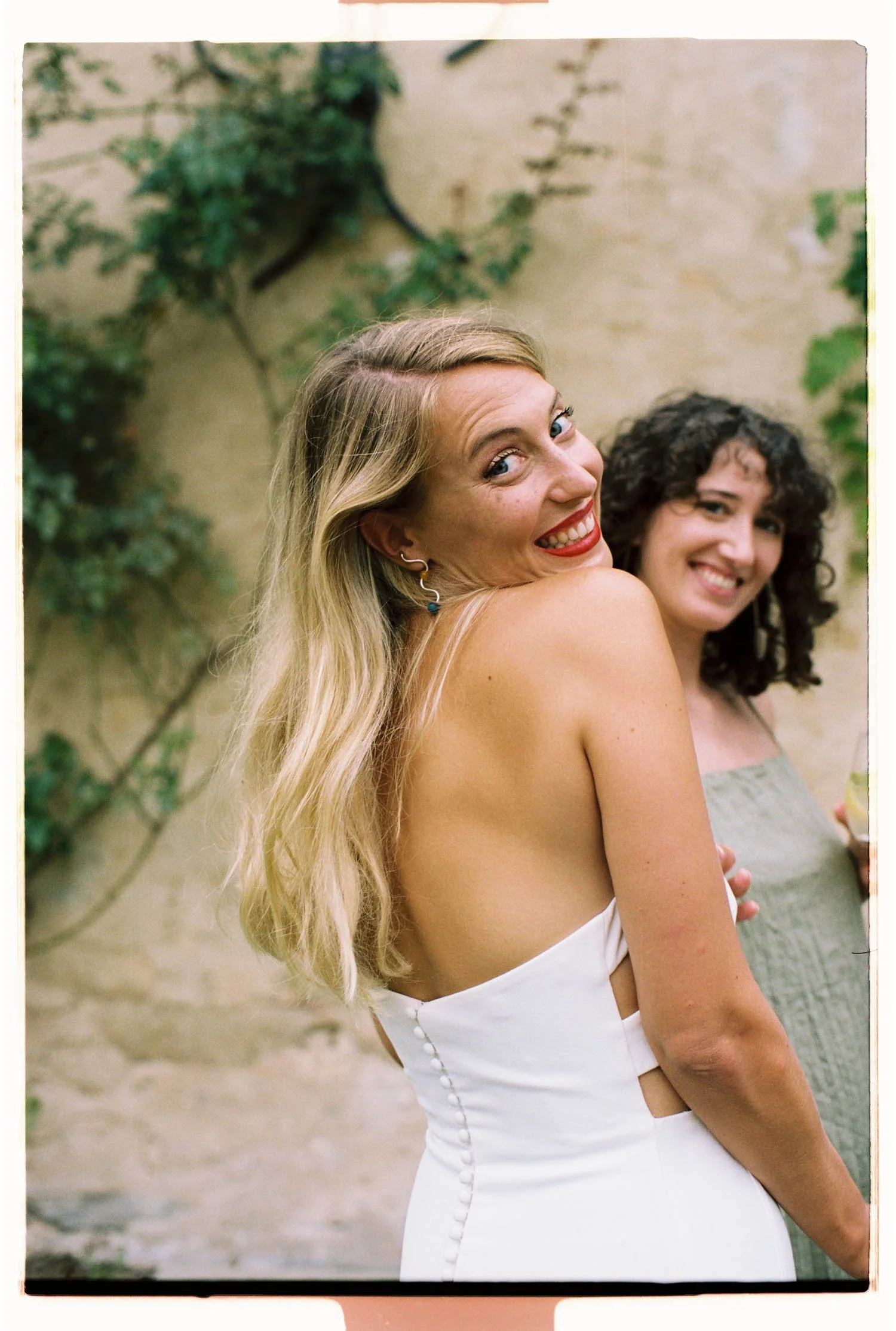 Candid analogue portrait of a bride smiling outdoors, standing against a beige wall with greenery.