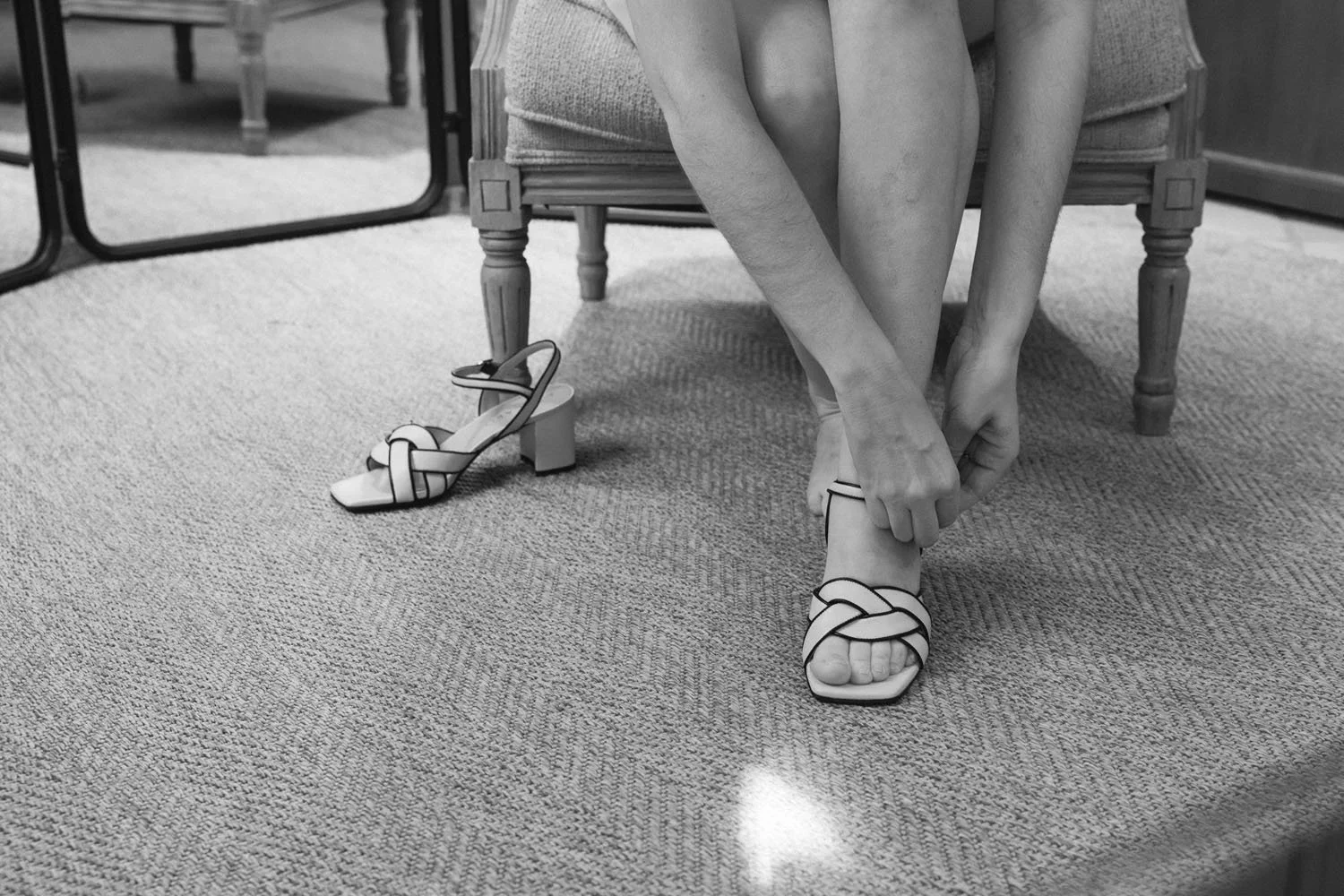 A woman is sitting on a wooden chair, putting on a high-heeled sandal. There is another matching sandal on the floor beside her, and she is wearing a similar pair on her feet. The room has a carpeted floor, a mirror, and some furniture.
