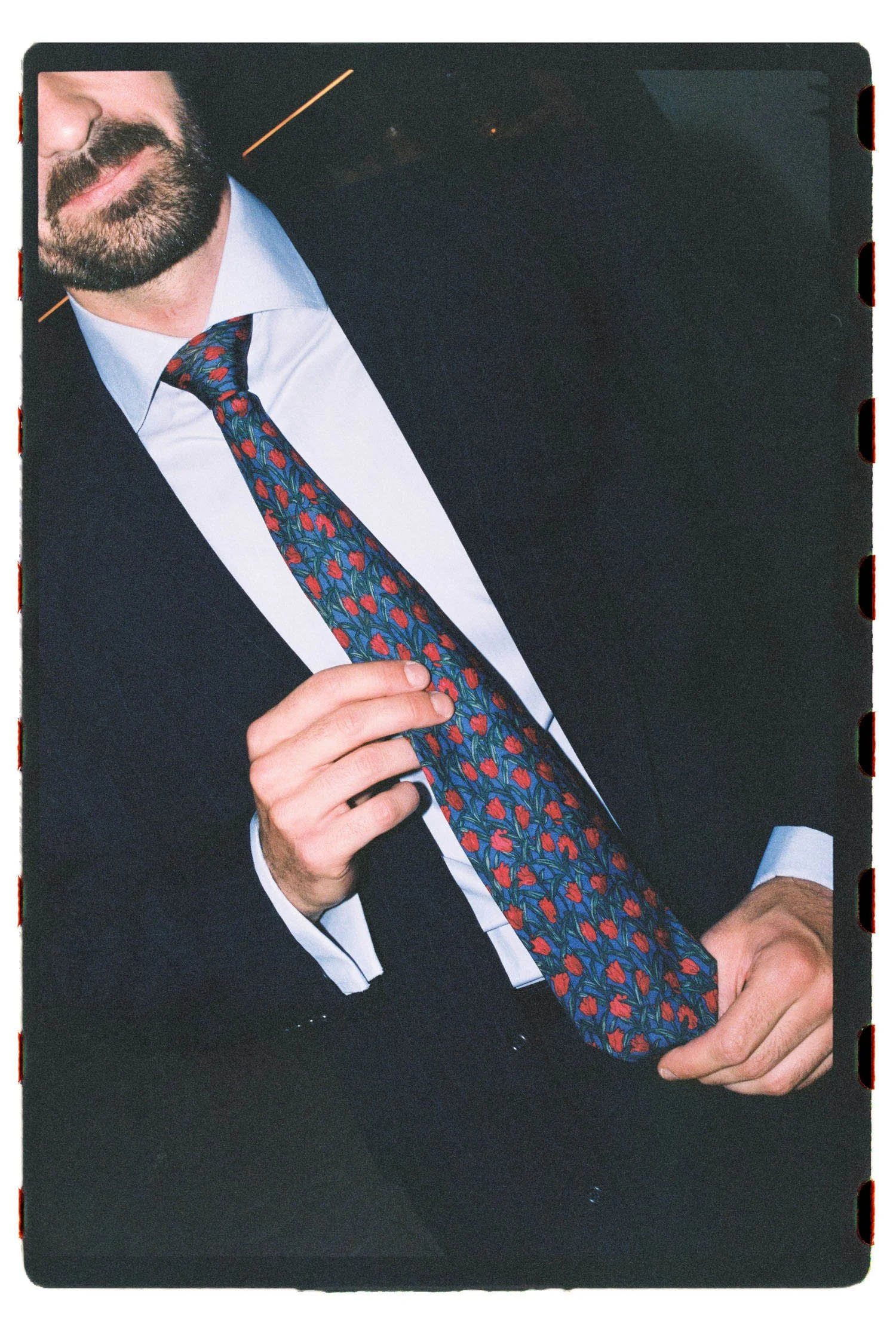 Close-up of a wedding guest in formal attire adjusting his colorful tie with a floral pattern. He has a well-groomed beard and is wearing a dark suit with a white shirt. Part of his face is visible, showing a subtle smile.