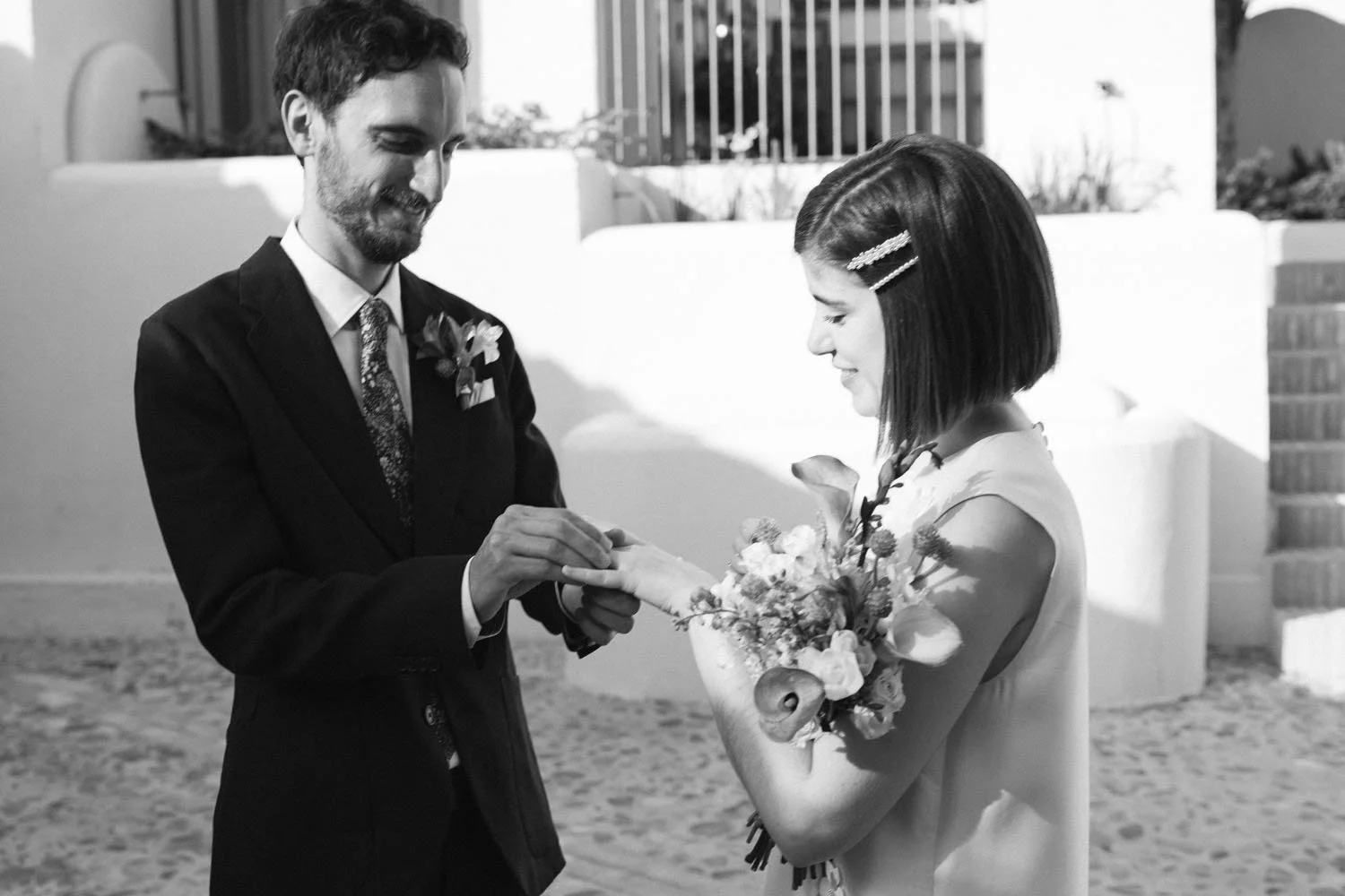 A black-and-white photo of a couple during a wedding ceremony. The groom is placing a ring on the bride's finger. The groom has dark hair and a beard, and is dressed in a suit with a floral boutonniere. The bride has short dark hair with hair clips, 