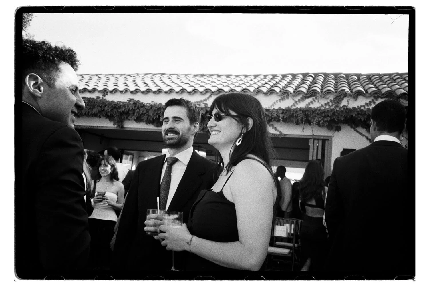 Black and white photo of a group of guests at a wedding outdoors, smiling and talking. A woman in the foreground is holding a drink, wearing sunglasses and earrings. Men around her are dressed in suits and are engaged in conversation.