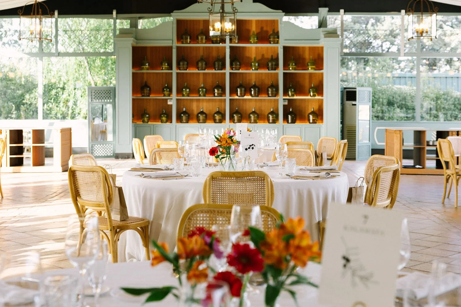 A decorated banquet hall with round tables covered in white tablecloths, set with glassware and silverware, and adorned with colorful flower centerpieces. Light streams in through large windows, revealing greenery outside. A large wooden shelving uni