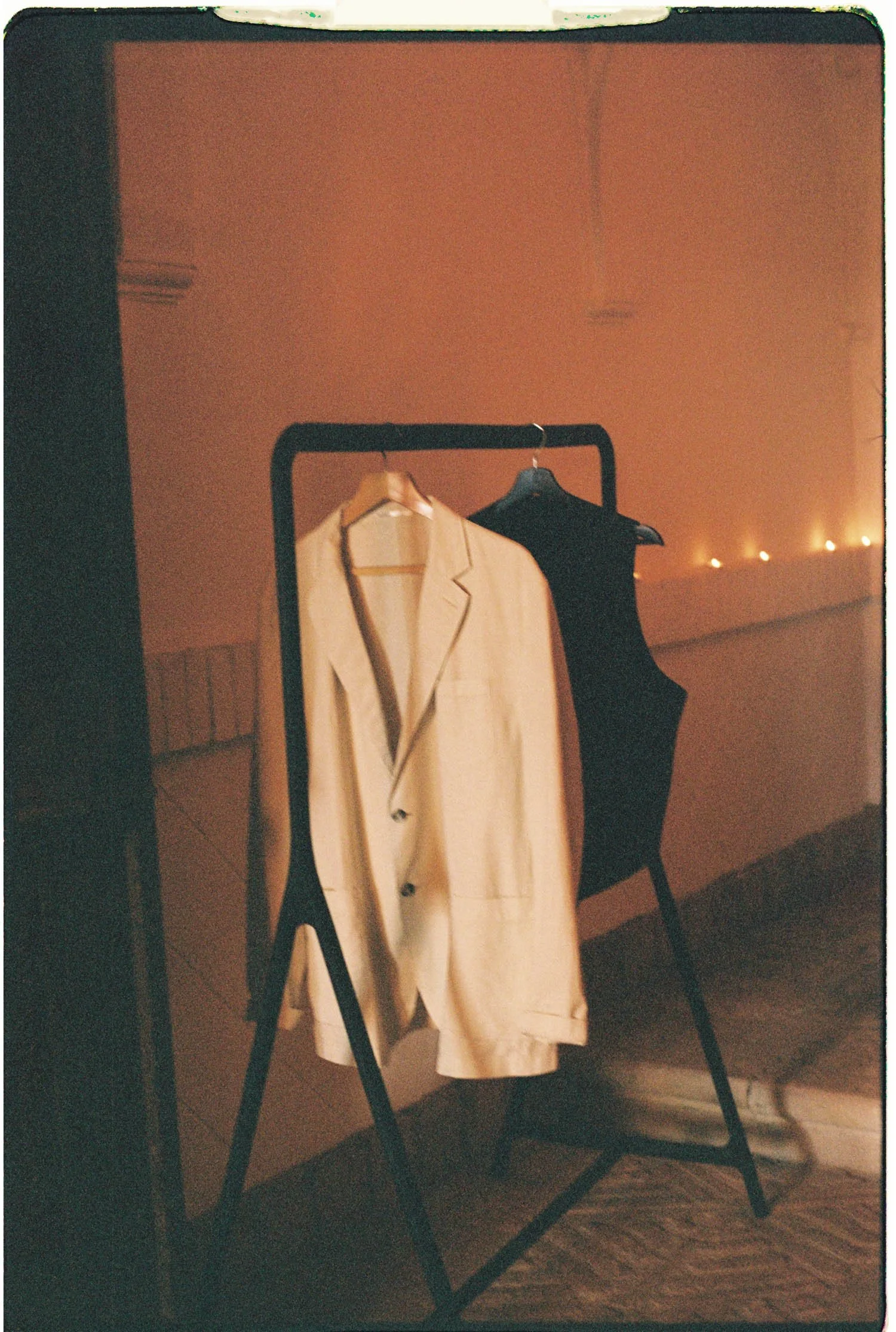 Black clothing rack holding a cream blazer and a black dress, set against a peach-colored wall with string lights in the background.