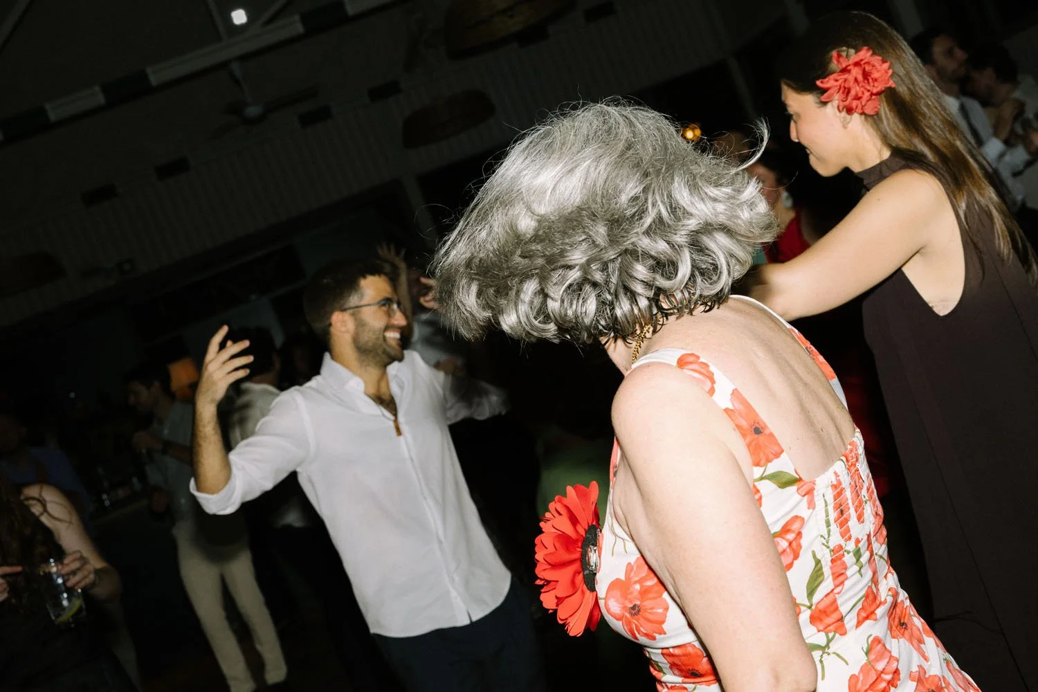 People dancing and socializing at a party, including a woman with curly gray hair wearing a floral dress, a young woman with long brown hair and a red flower hair accessory, and a man in a white shirt smiling and raising his hand.