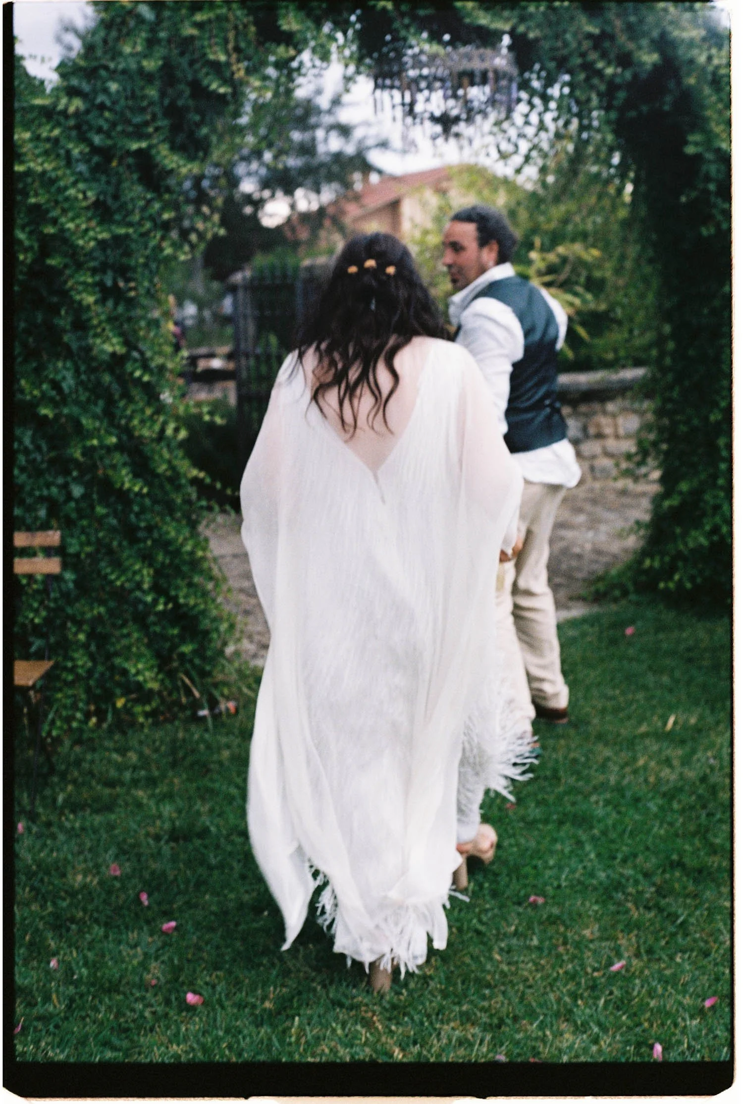 Analogue candid photo of a bride in a white dress walking on grass, with groom in a vest and white shirt looking at her, surrounded by greenery and pink flower petals.