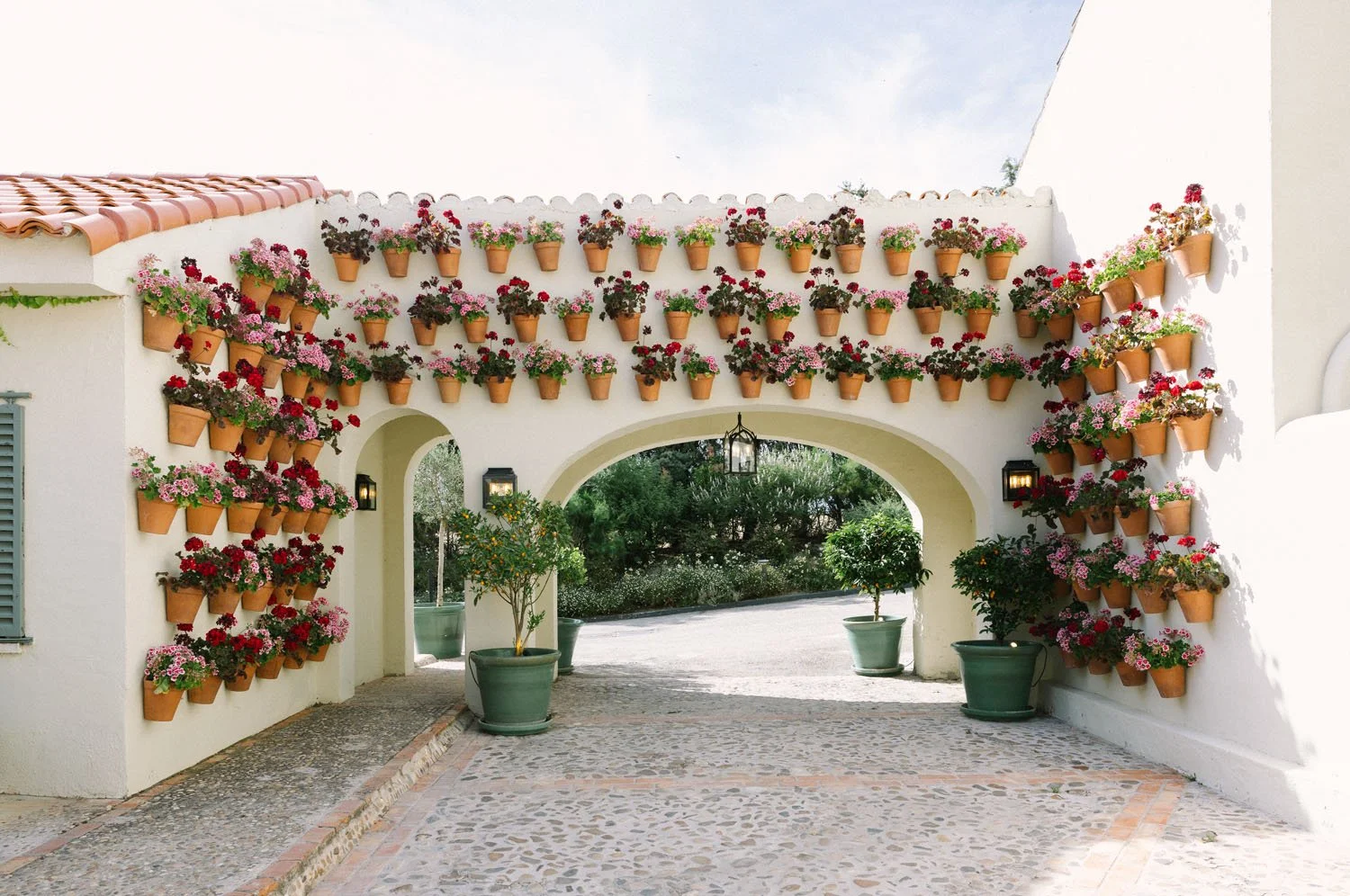 Archway with white walls decorated with terracotta pots filled with pink and red flowering plants, flanked by potted plants, leading to a garden.