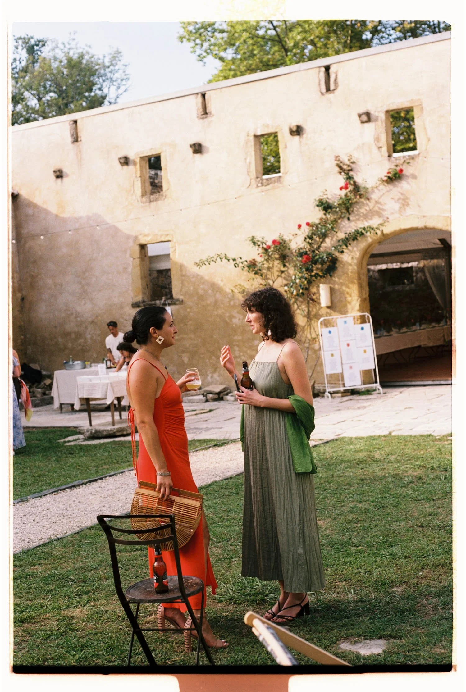 Two women are outside at a social gathering, engaged in conversation. One is wearing a red dress, holding a drink and a woven handbag, with high heels. The other is dressed in a long greenish-gray dress with a green sweater draped over her shoulders 