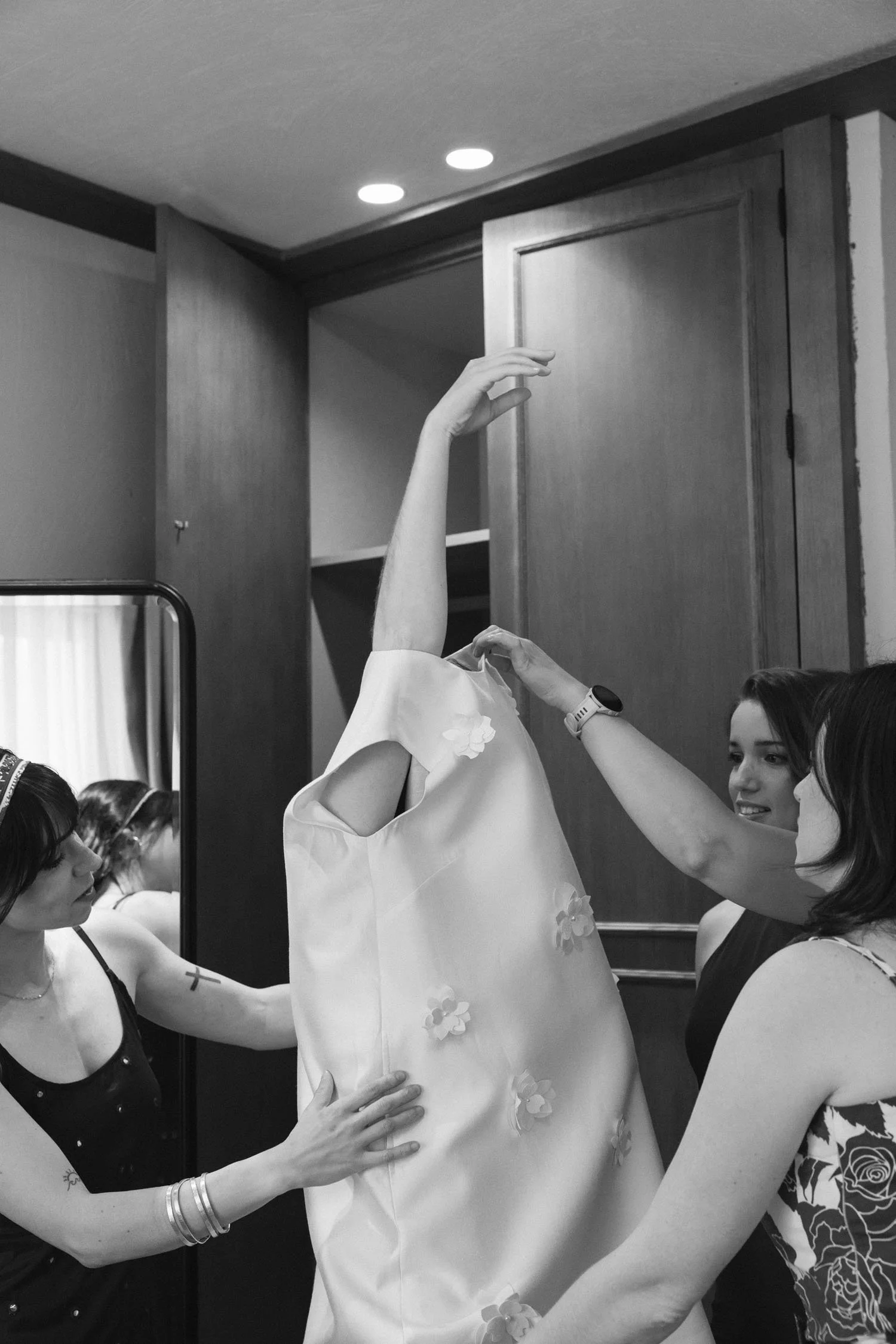 Several women are helping a bride get ready, adjusting her dress, in a room with wood-paneled walls and ceiling lights.