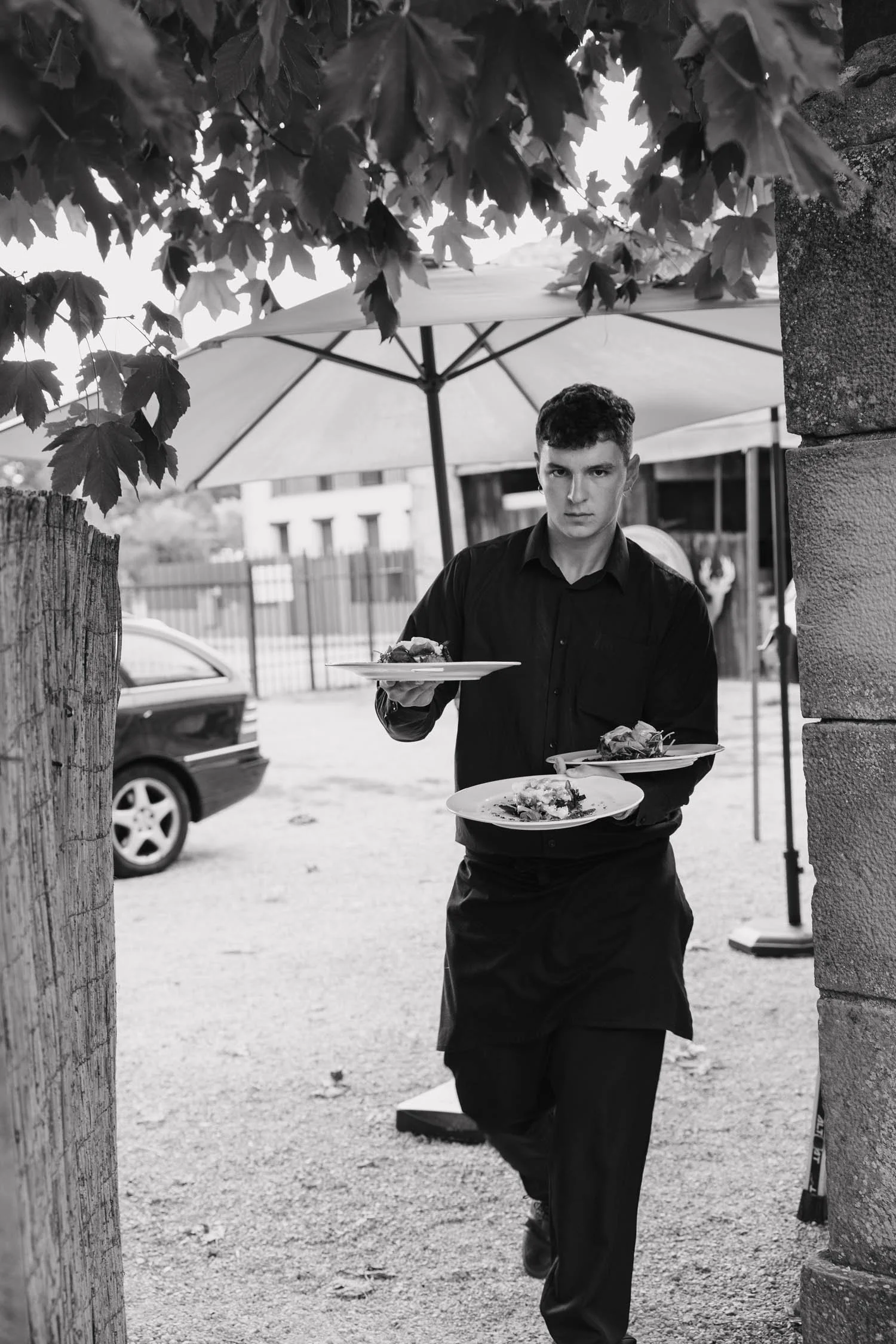 A young man in black clothes serving plates of food outdoors in a wedding under a large umbrella, with trees, a car, and buildings in the background.