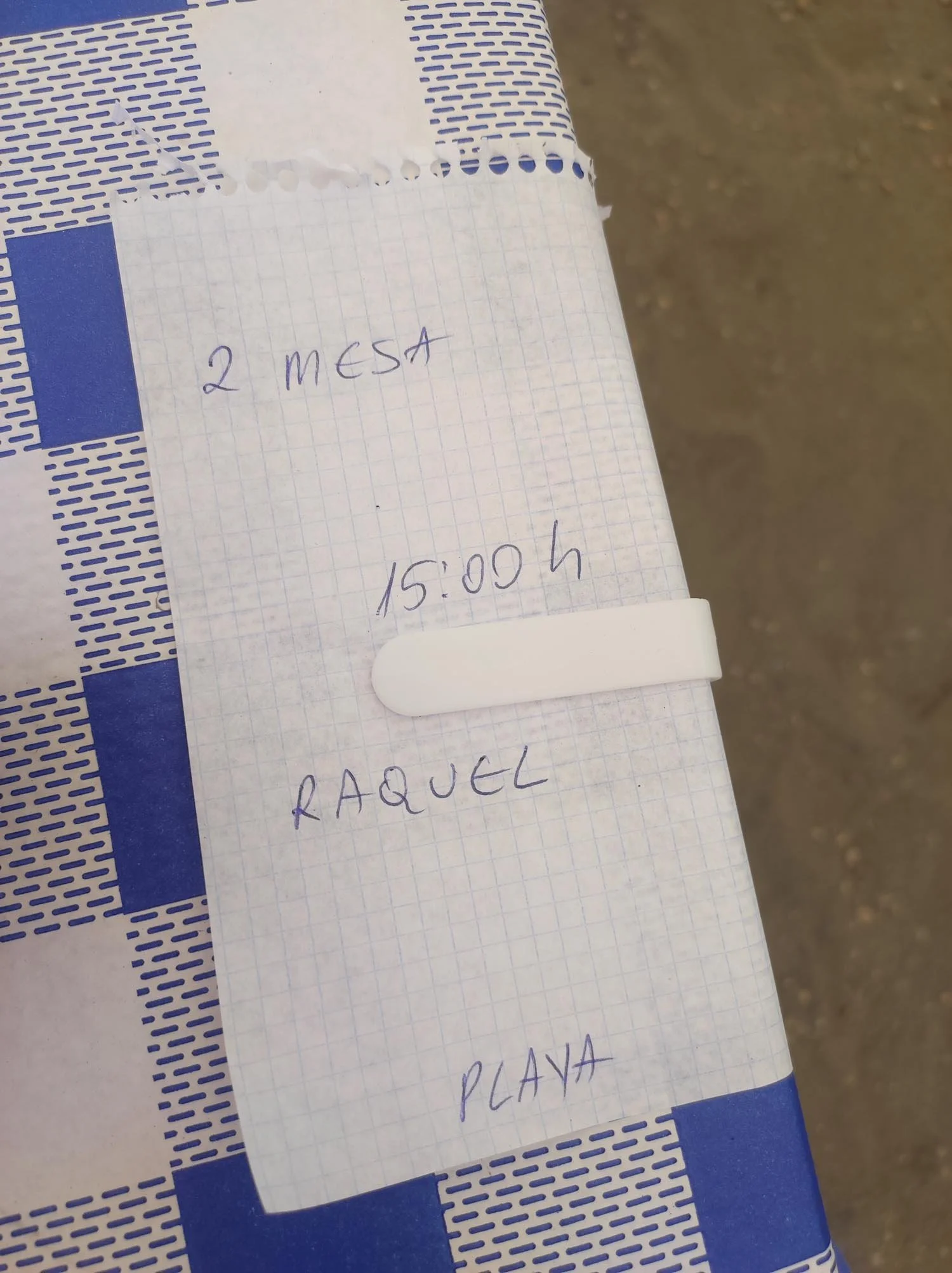 A handwritten note on graph paper with blue ink that reads '2 MESA' at the top, a time '15:00', and part of another word 'PARA' with additional text not fully visible. The paper is on a blue and white patterned surface, with part of a dirt ground visible in the background.