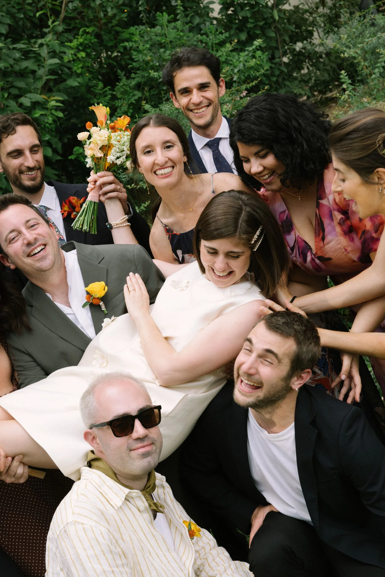 A group of people at a wedding or celebration gathered closely together outdoors, with greenery in the background. They are smiling, and a woman in a white dress is lying on top of the group. Some are holding flowers, and everyone appears joyful and 