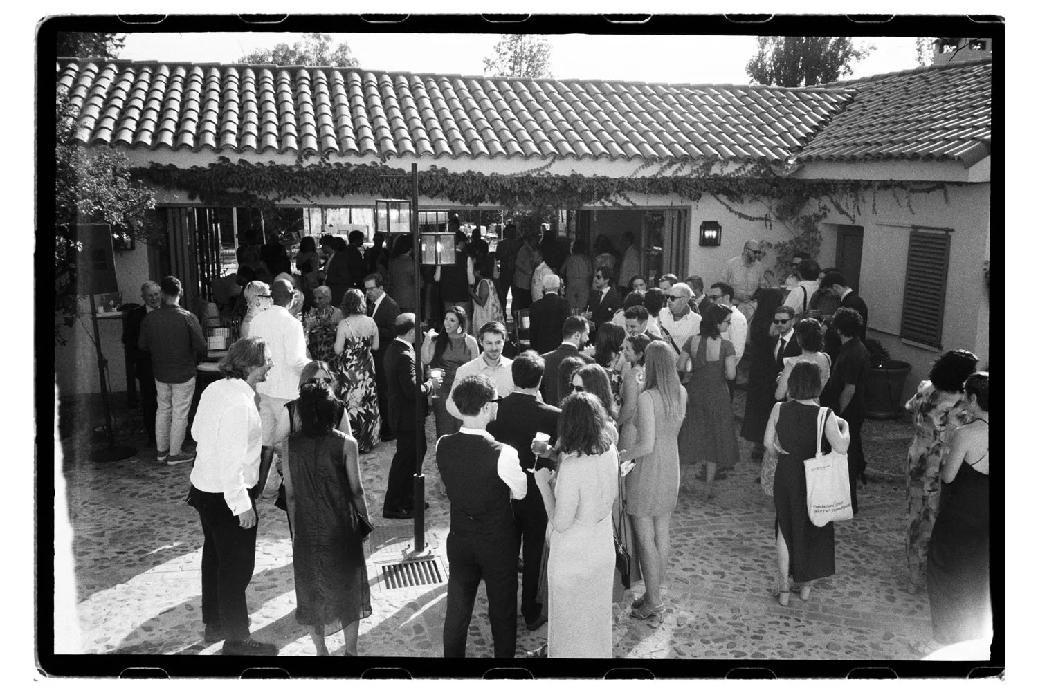 A black-and-white photo of wedding guests outside a building with a tiled roof. People are mingling, chatting, and holding drinks. The scene is lively with a mix of men and women dressed in semi-formal and casual attire.